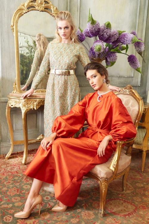 Two women model dresses by Anjilla Seddeqi in a room styled with gilded furniture and flowers.