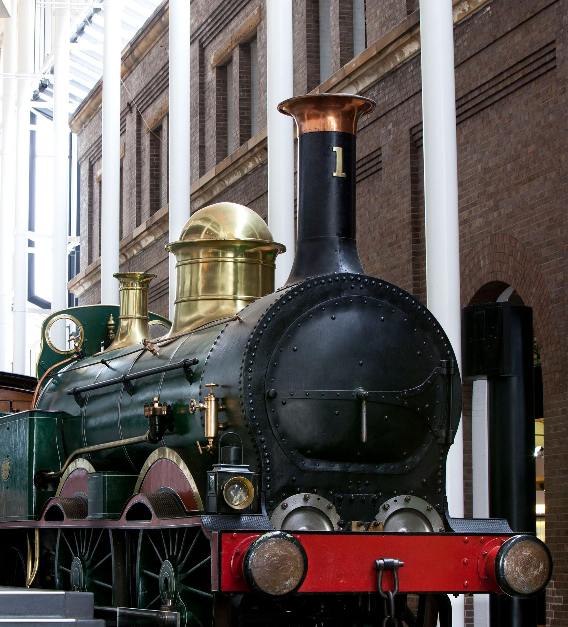 The front of a large steam engine with a red front bar, black body and gold cylinders.