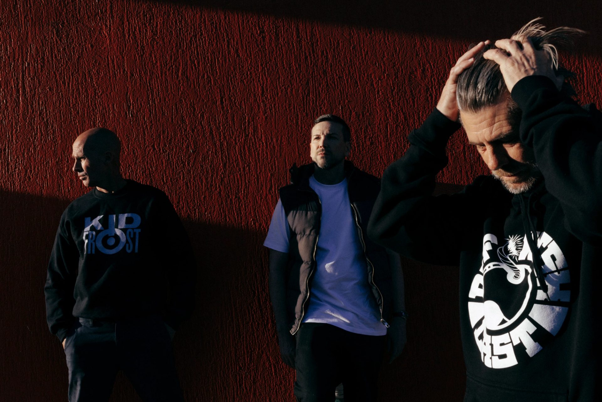 Thee men standing near a maroon wall.