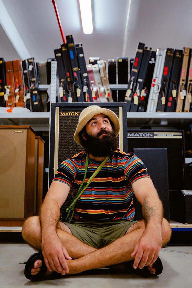 Joseph Douglas sits crossed legged on the floor in front of a row of guitar amps. In the background is a shelf lined with guitar cases.