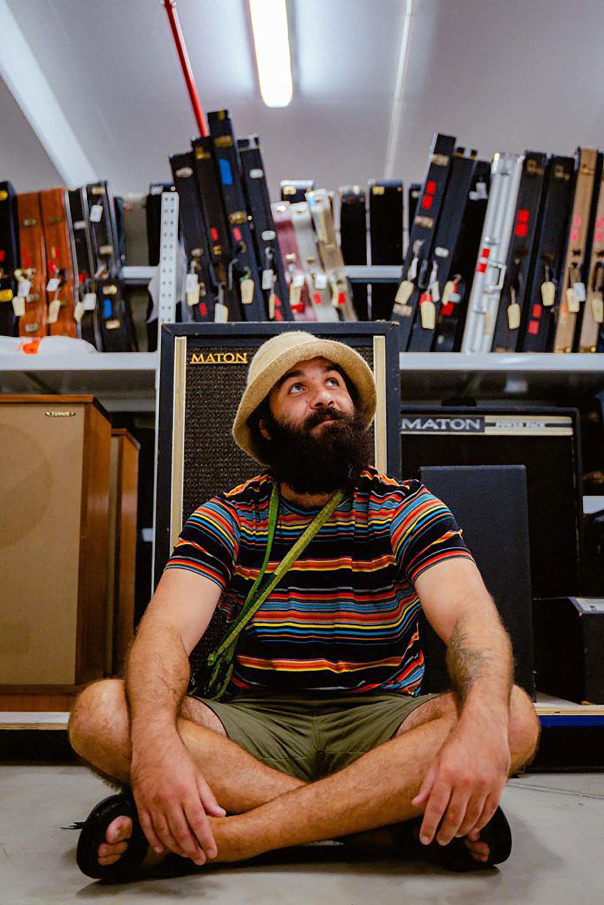 Joseph Douglas sits crossed legged on the floor in front of a row of guitar amps. In the background is a shelf lined with guitar cases.
