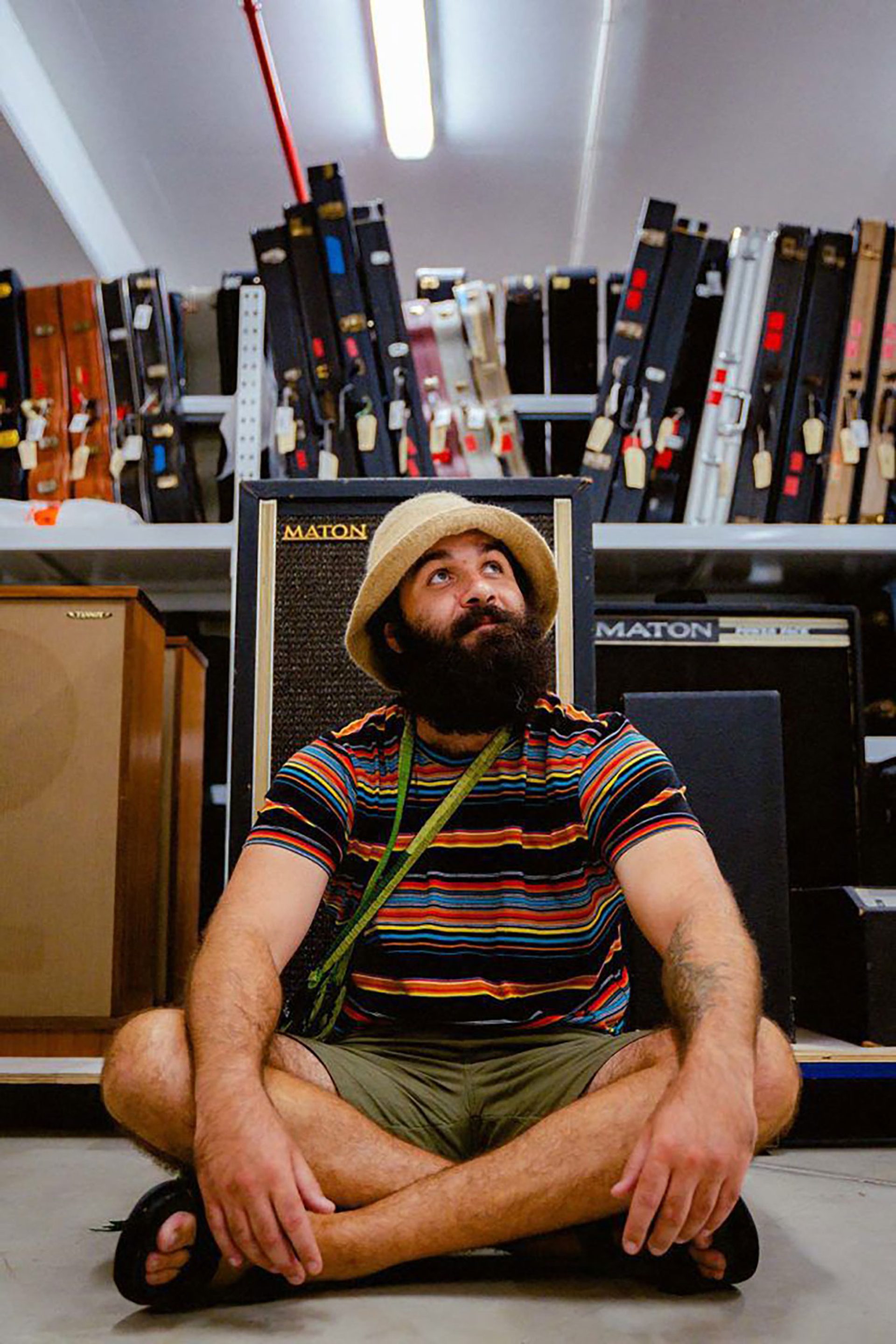 Joseph Douglas sits crossed legged on the floor in front of a row of guitar amps. In the background is a shelf lined with guitar cases.
