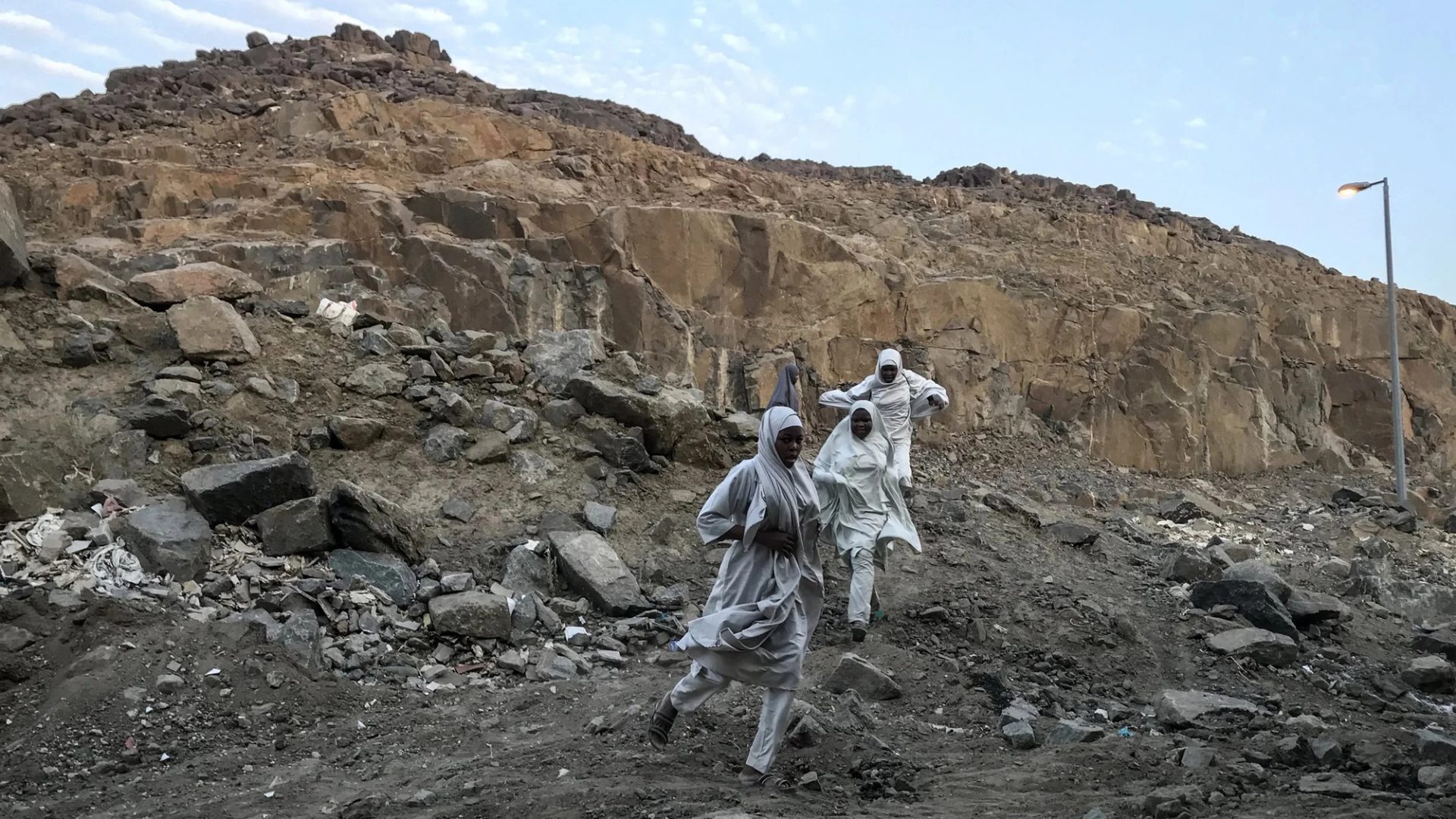 A photograph of three figures in a large rock quarry, wearing white robes, running towards the camera. The sky behind them is bright blue.