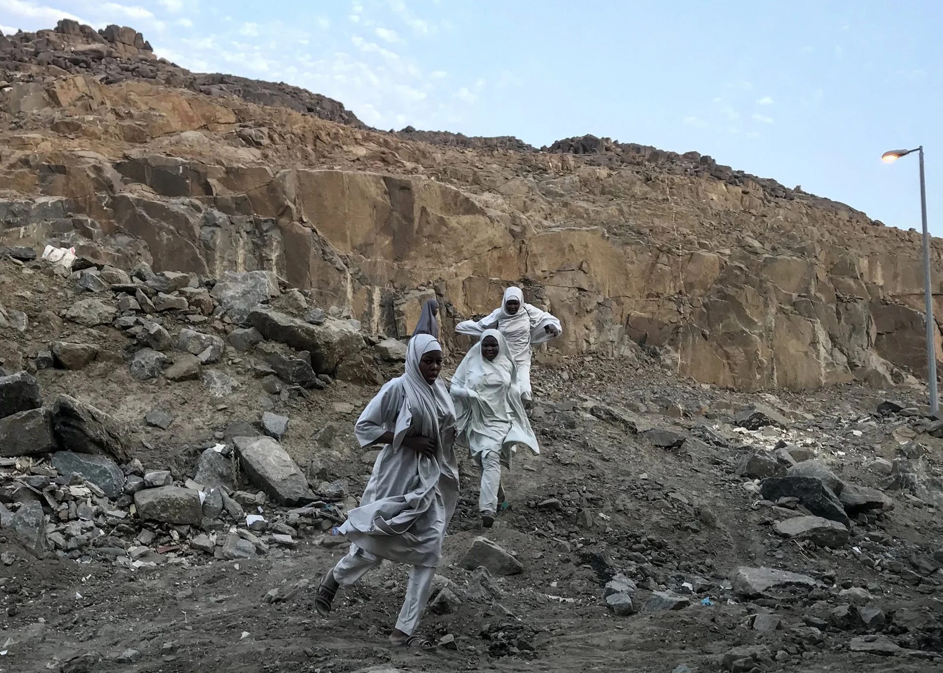A photograph of three figures in a large rock quarry, wearing white robes, running towards the camera. The sky behind them is bright blue.