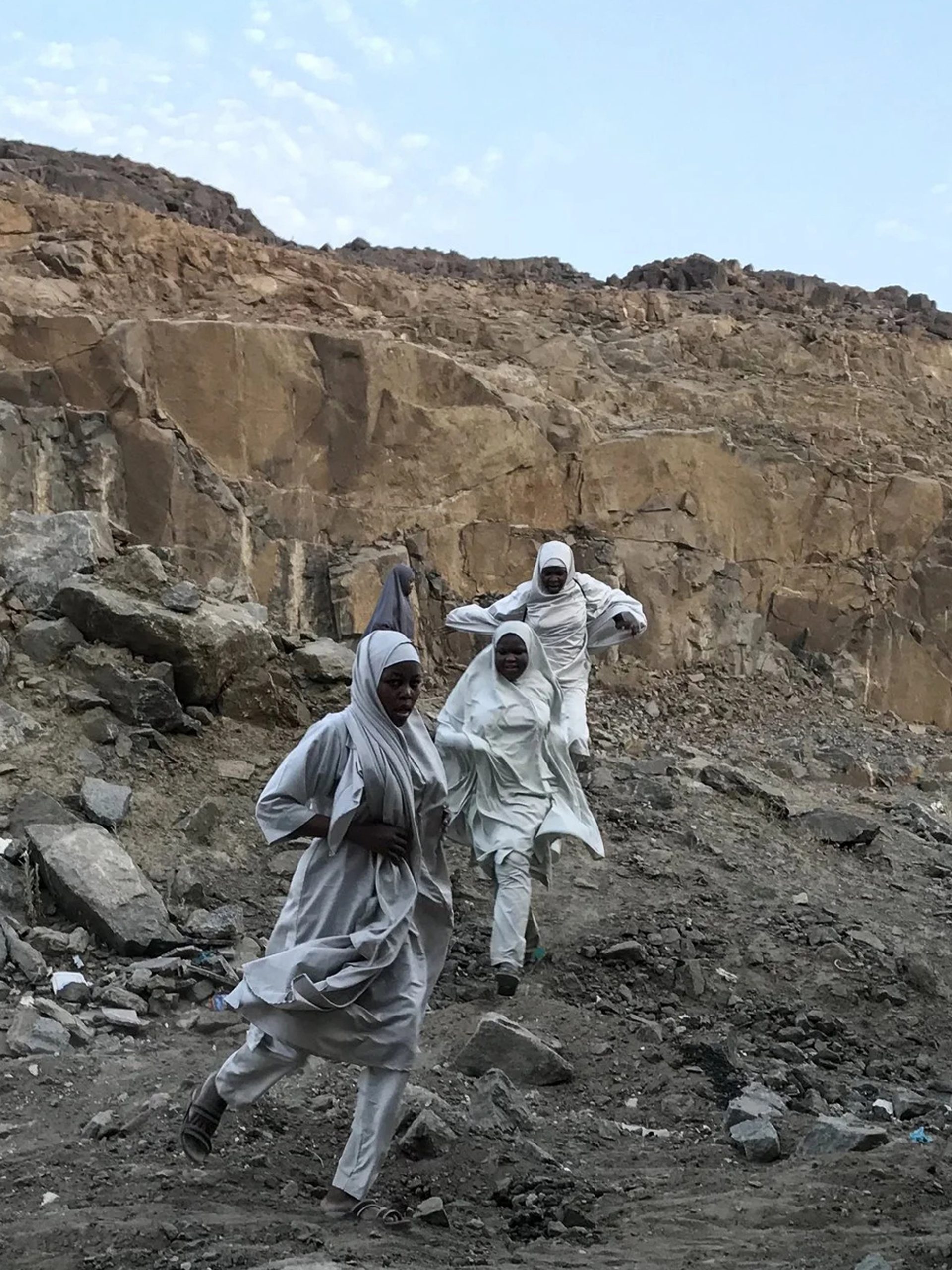 A photograph of three figures in a large rock quarry, wearing white robes, running towards the camera. The sky behind them is bright blue.