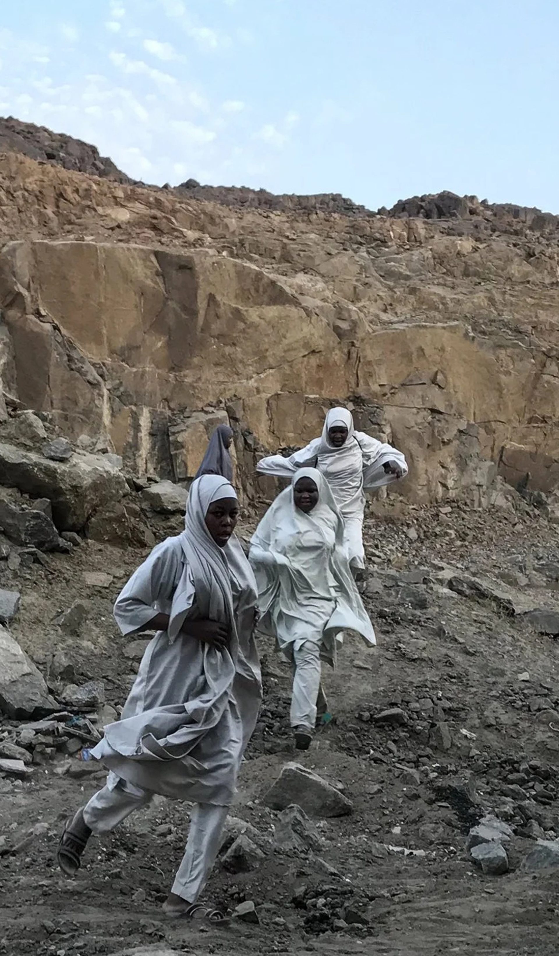 A photograph of three figures in a large rock quarry, wearing white robes, running towards the camera. The sky behind them is bright blue.