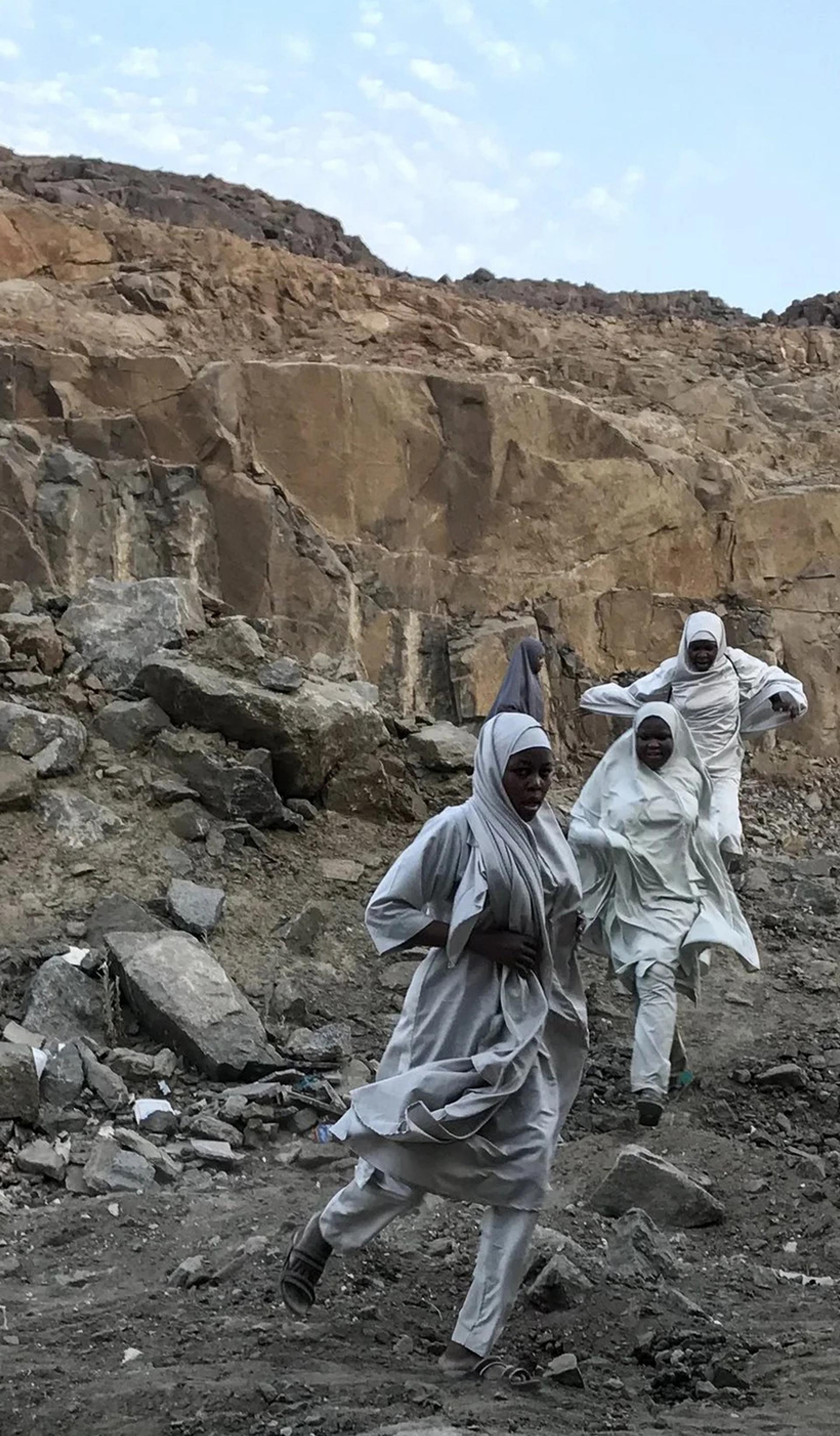 A photograph of three figures in a large rock quarry, wearing white robes, running towards the camera. The sky behind them is bright blue.
