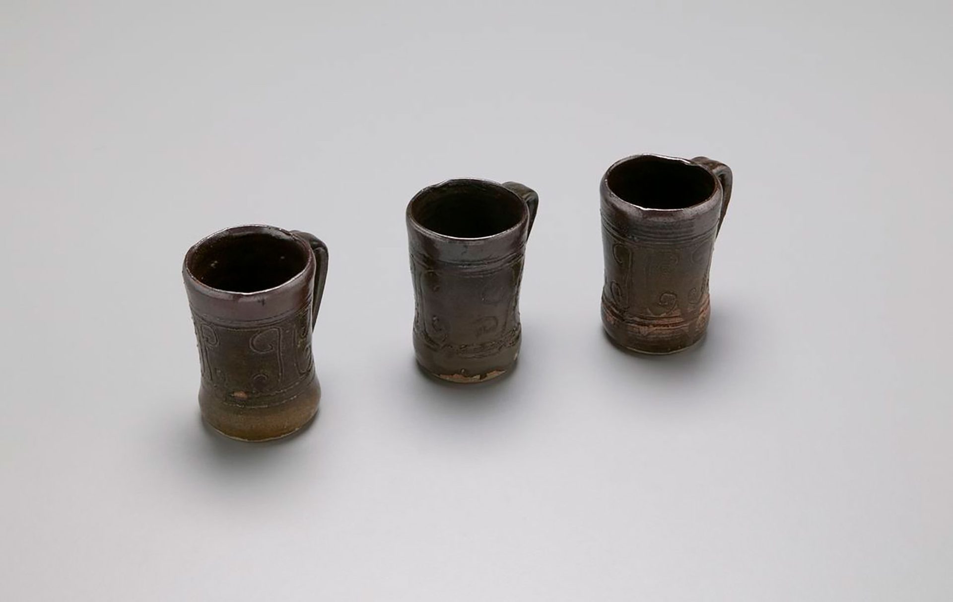 Three mugs with handles with a tenmoku glaze. Inscribed with simple curved lines in an abstract design engraved on the mugs.