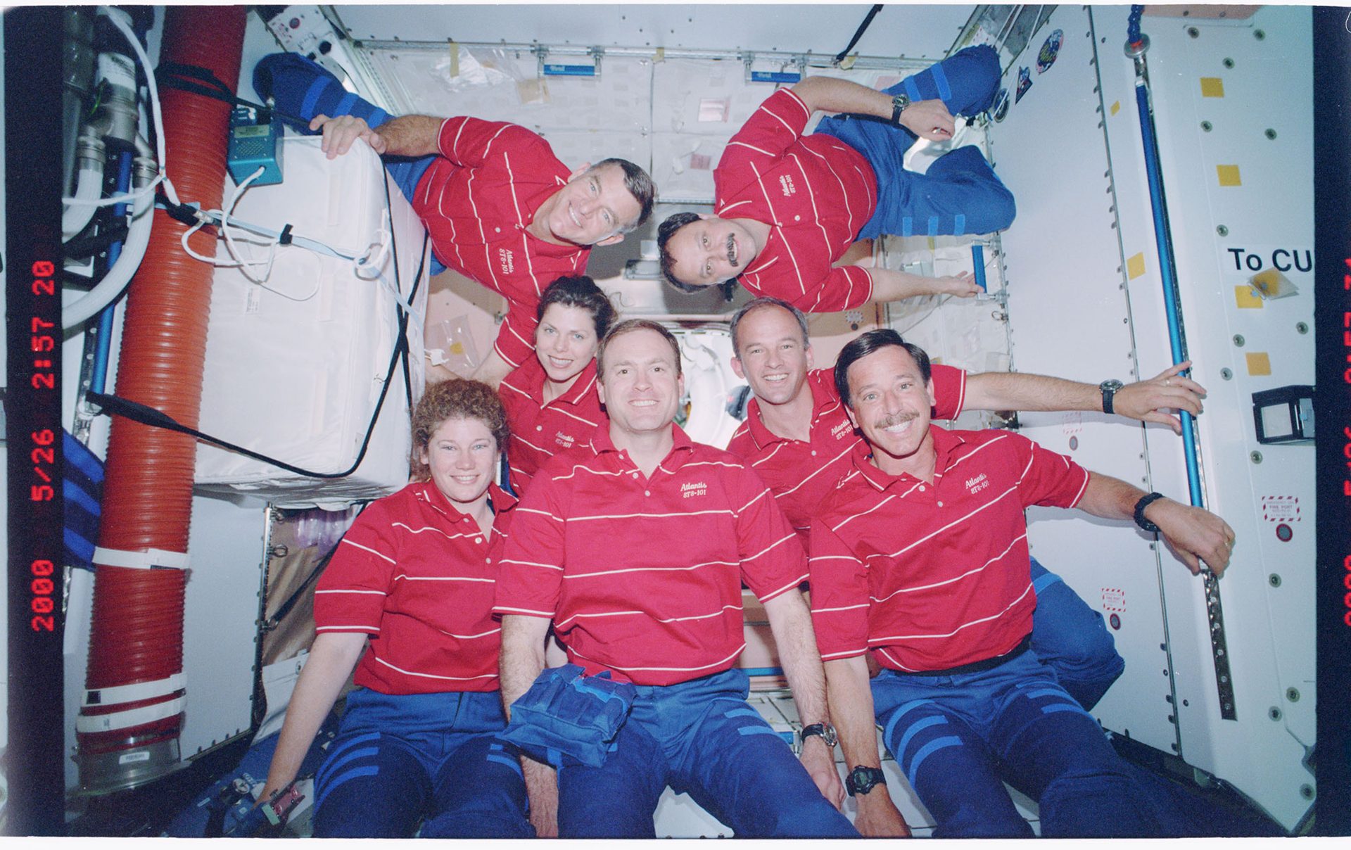 Seven people wearing red striped polo shorts and blue trousers float inside a space capsule