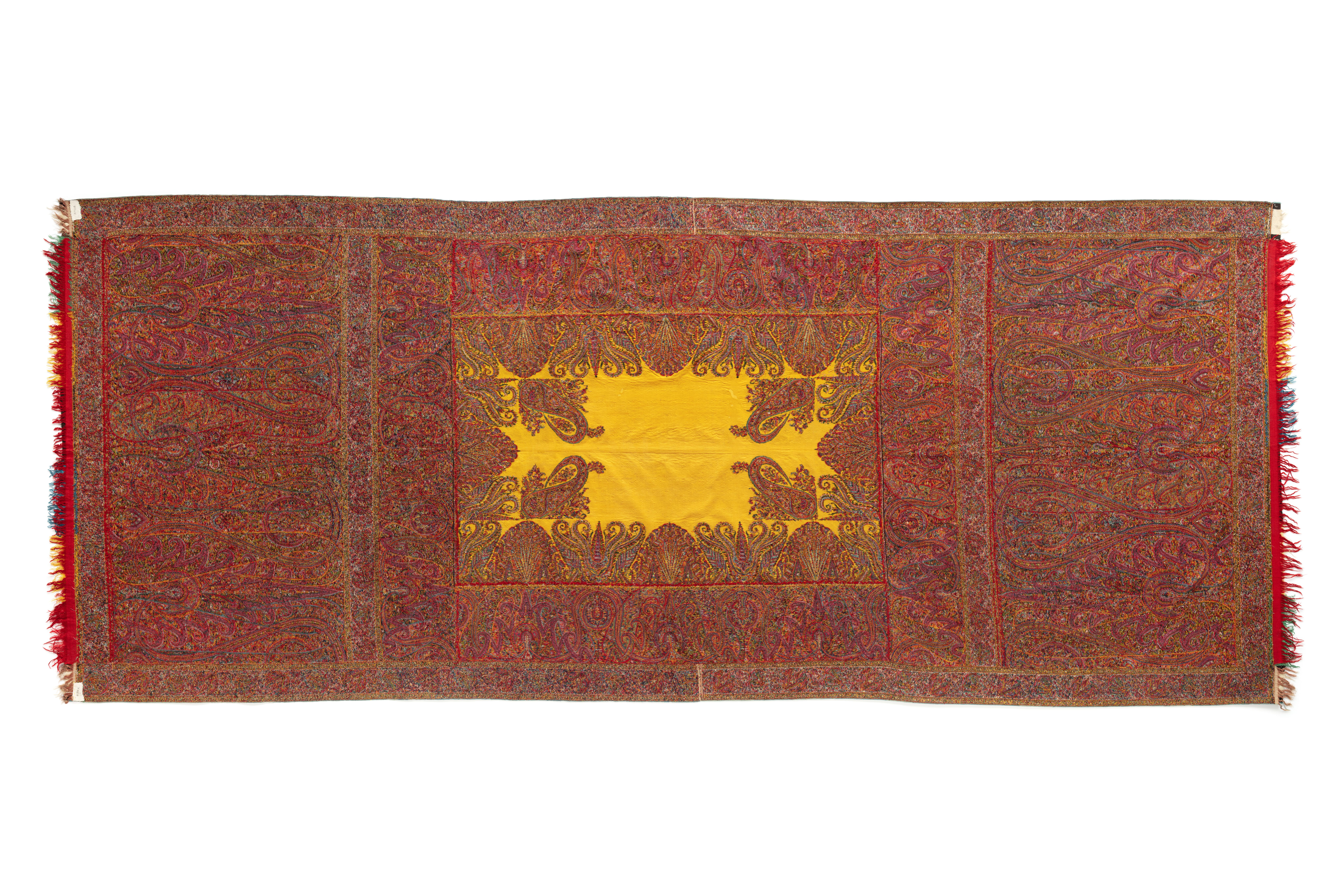 A rectangular Kashmiri long shawl with a rectangular yellow centre surrounded by wide side borders and deep end borders. The borders are intricately and very finely patterned with a complex design of elongated boteh motifs; the design also encroaches delicately into the yellow field. Using the kani technique, the shawl has been woven in sections which were then hand stitched together to form the whole. Each end is finished with a narrow row of fringed patchwork pieces of red, blue, green, yellow, white and black fabric.