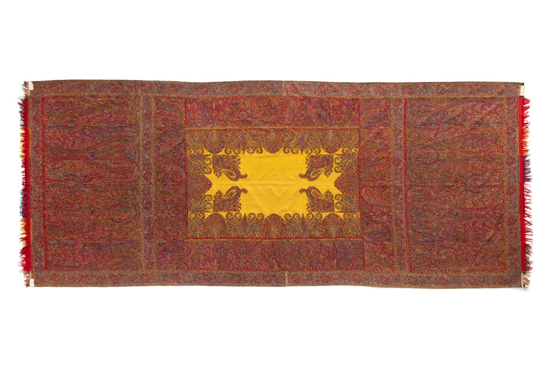 A rectangular Kashmiri long shawl with a rectangular yellow centre surrounded by wide side borders and deep end borders. The borders are intricately and very finely patterned with a complex design of elongated boteh motifs; the design also encroaches delicately into the yellow field. Using the kani technique, the shawl has been woven in sections which were then hand stitched together to form the whole. Each end is finished with a narrow row of fringed patchwork pieces of red, blue, green, yellow, white and black fabric.