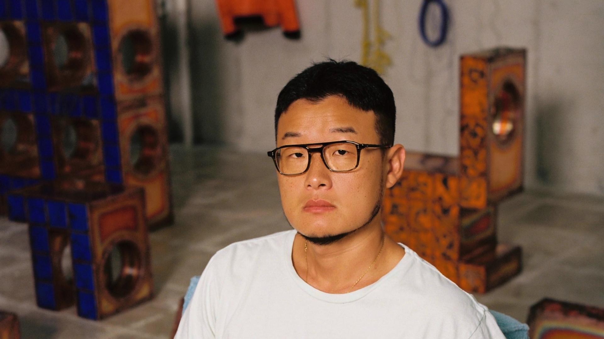 A man in a white t-shirt, beige trousers and black glasses looks into the camera.