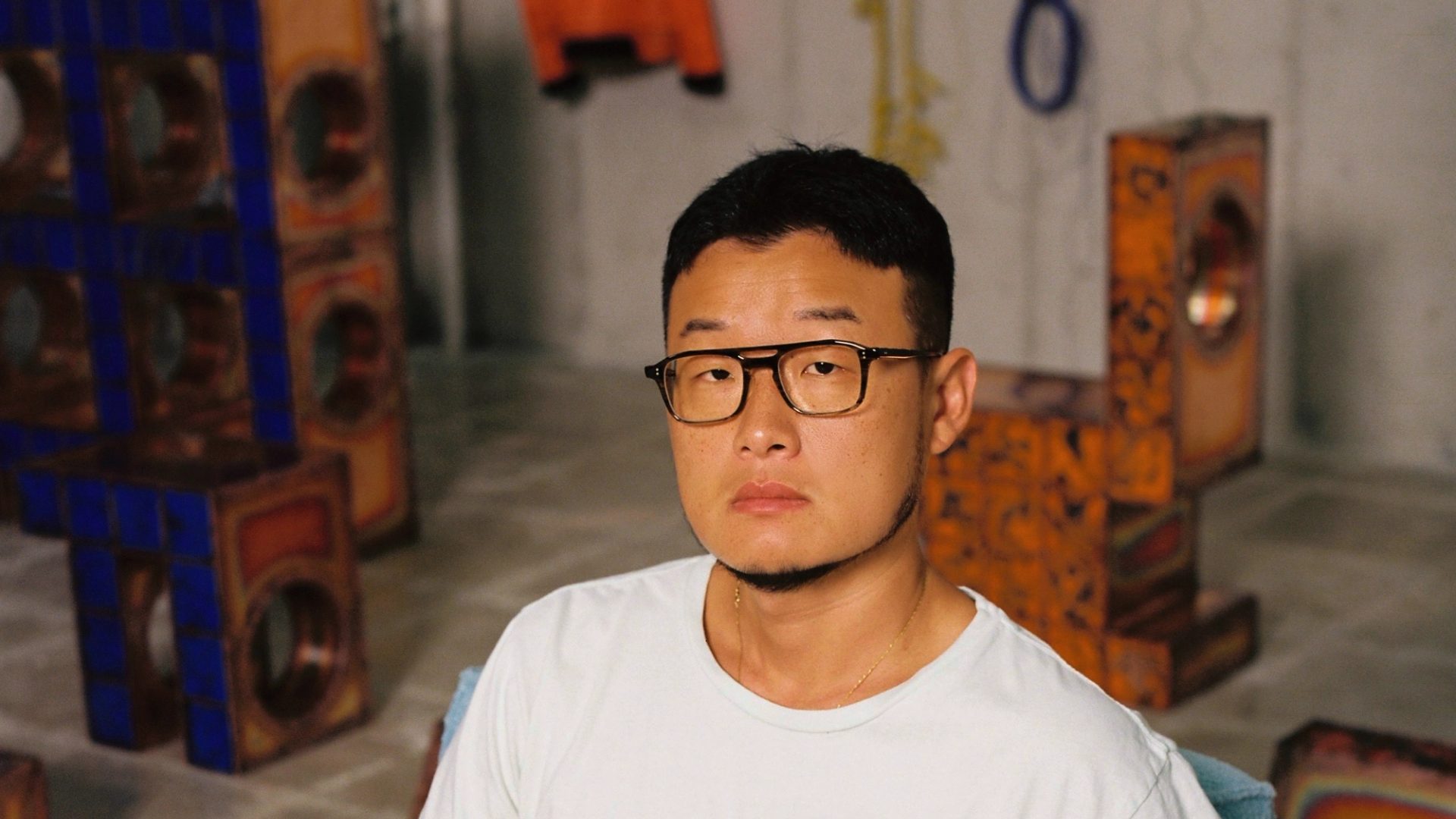 A man in a white t-shirt, beige trousers and black glasses looks into the camera.