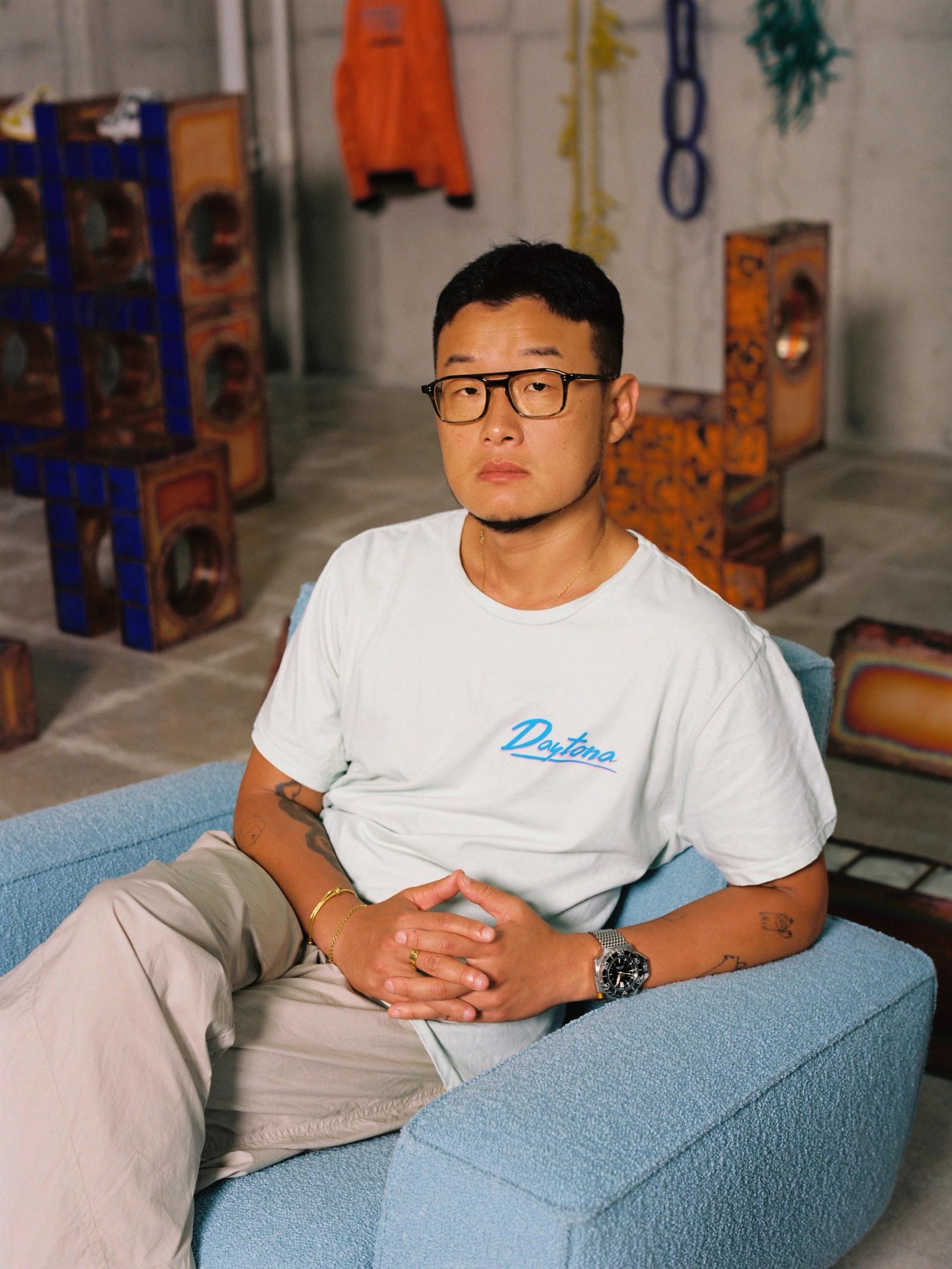 A man in a white t-shirt, beige trousers and black glasses looks into the camera.