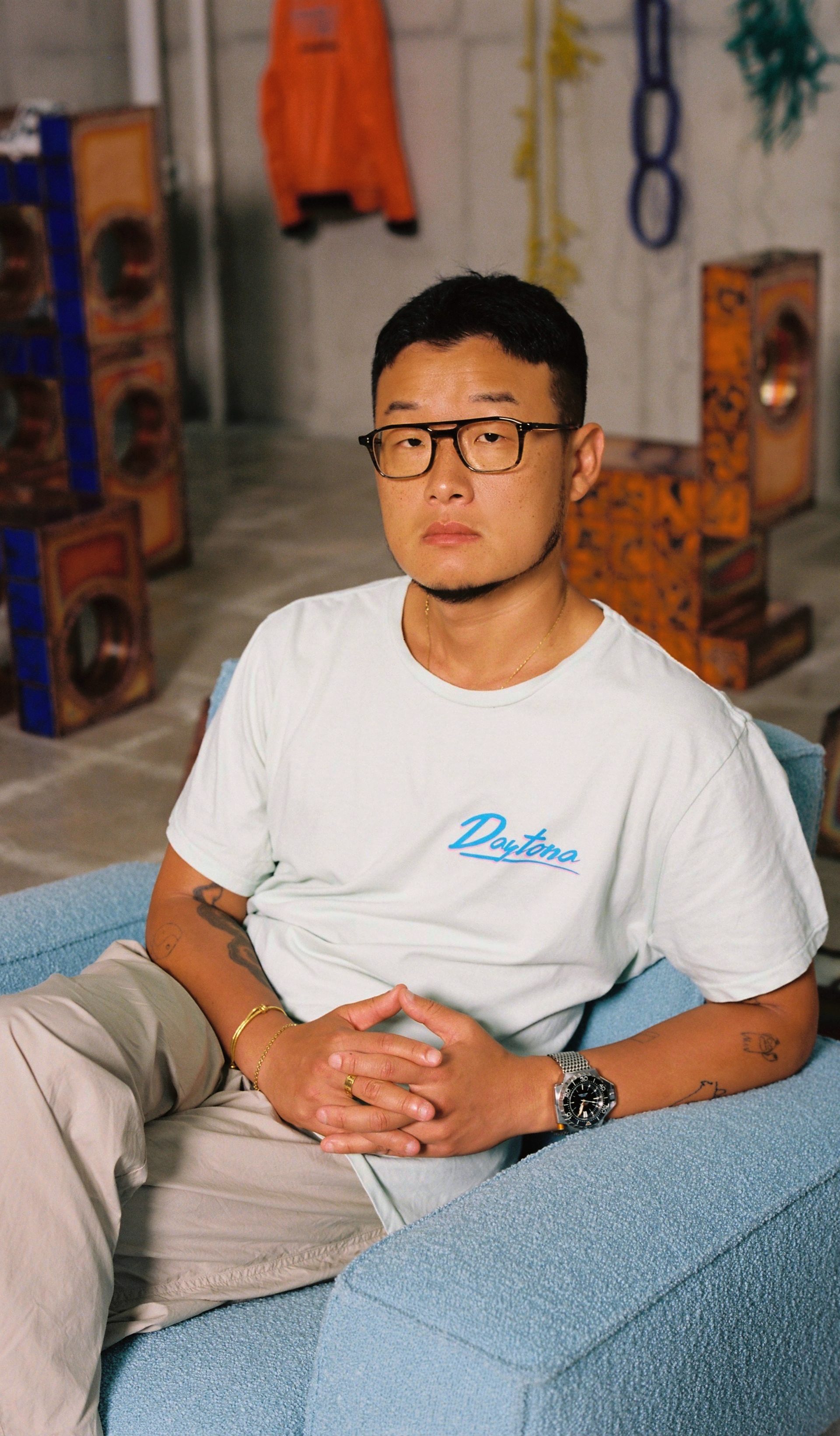 A man in a white t-shirt, beige trousers and black glasses looks into the camera.