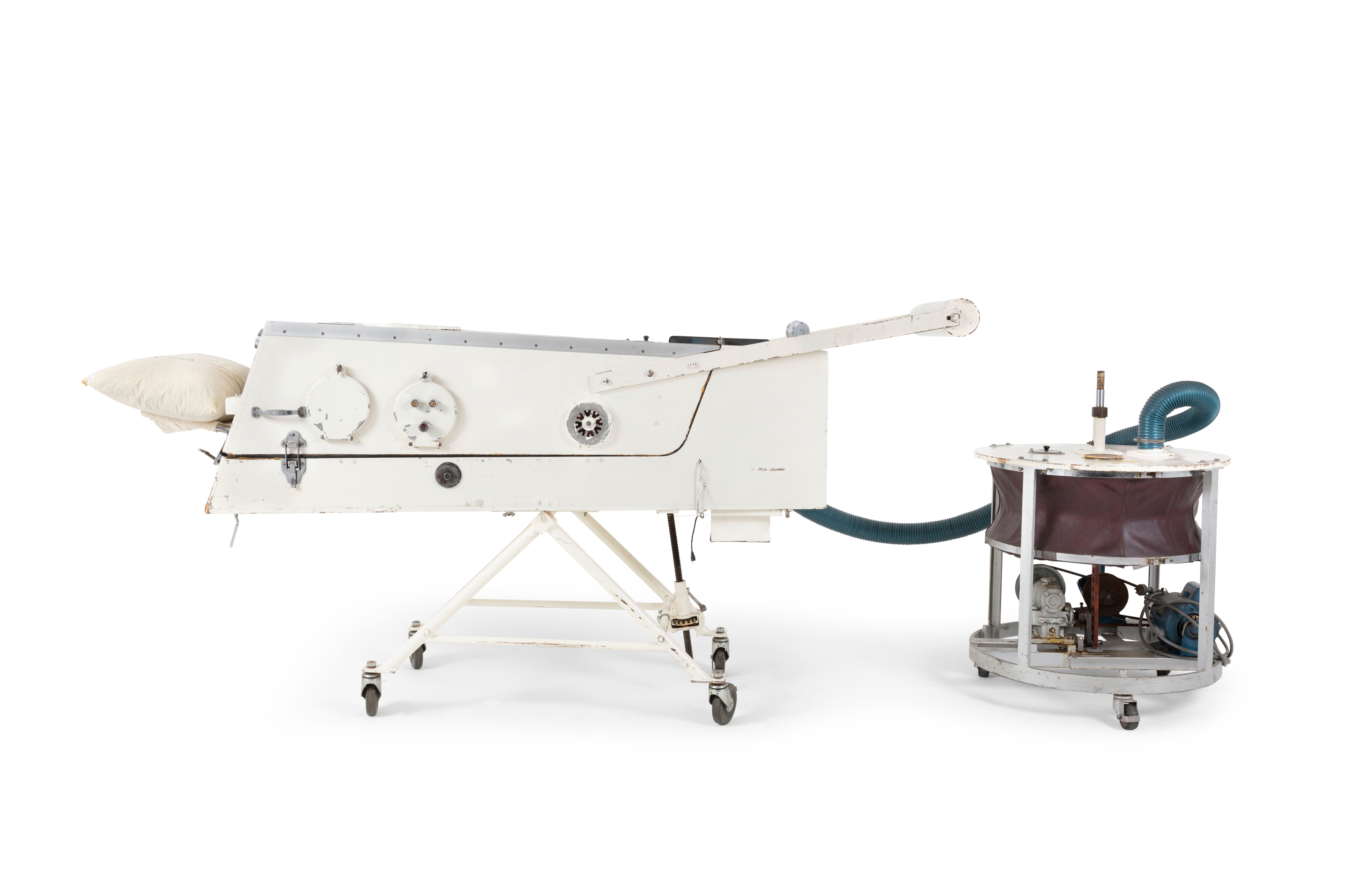 An artificial respirator, made of wood, metal and rubber. It shows a white painted cabinet standing on metal frame support with wheels, attached by a blue, flexible, pleated tube to electrical air pump on stand, with three castors; cabinet has plaque with wording, 'This respirator was given to 'Cherrywood' by the Elamang Auxiliary of the Poliomyelitis Society 24th August 1955'.