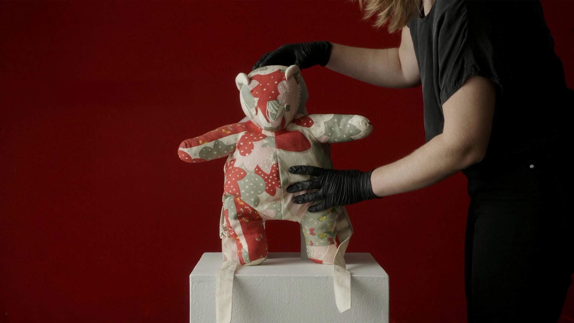A patchwork teddy bear held by a museum curator against a red background.