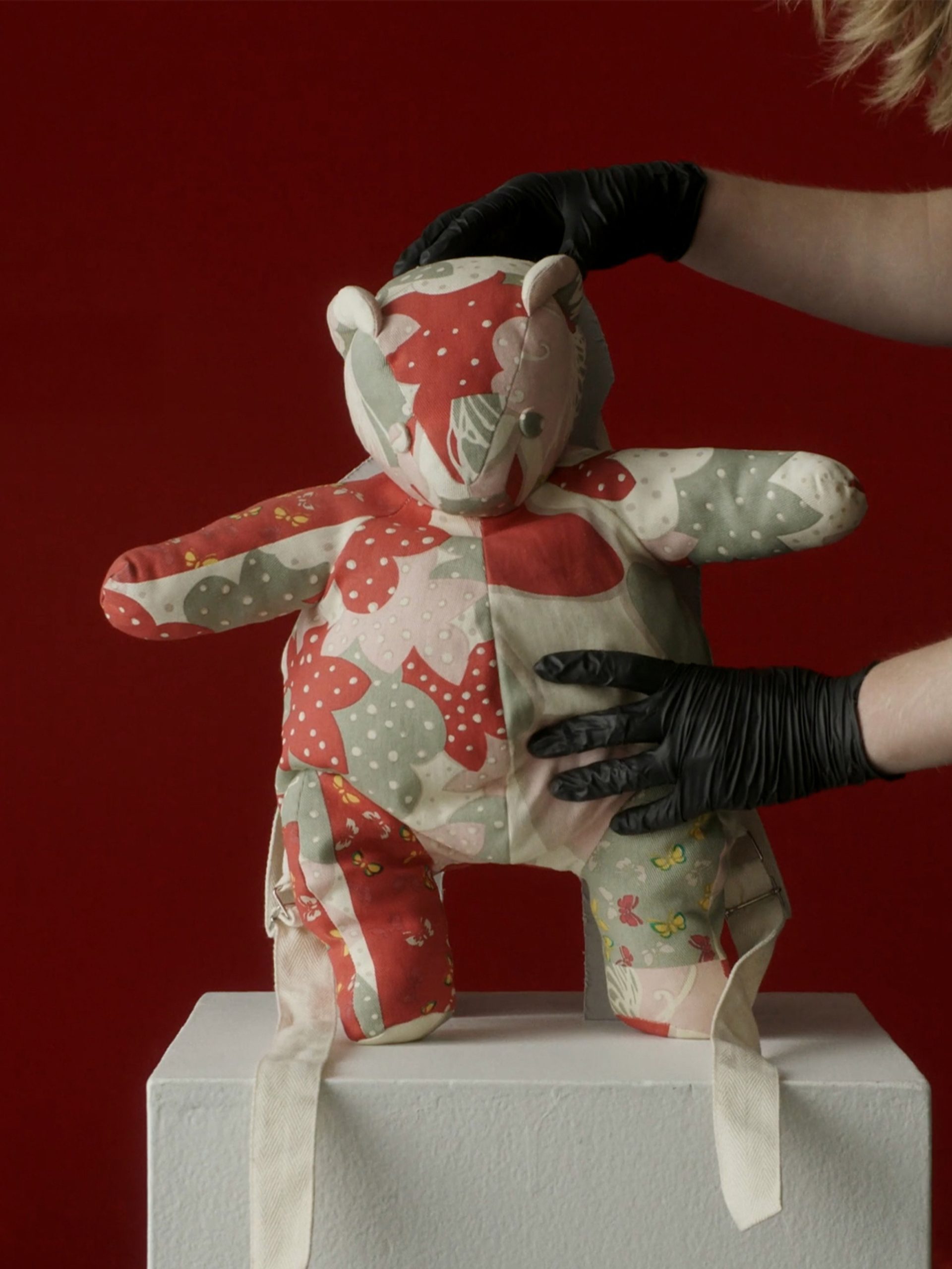A patchwork teddy bear held by a museum curator against a red background.