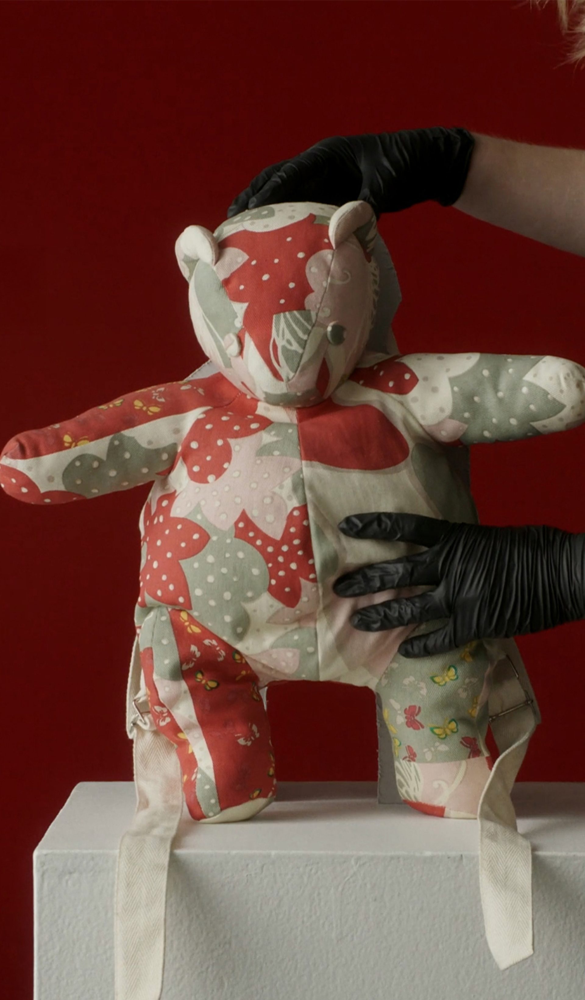 A patchwork teddy bear held by a museum curator against a red background.