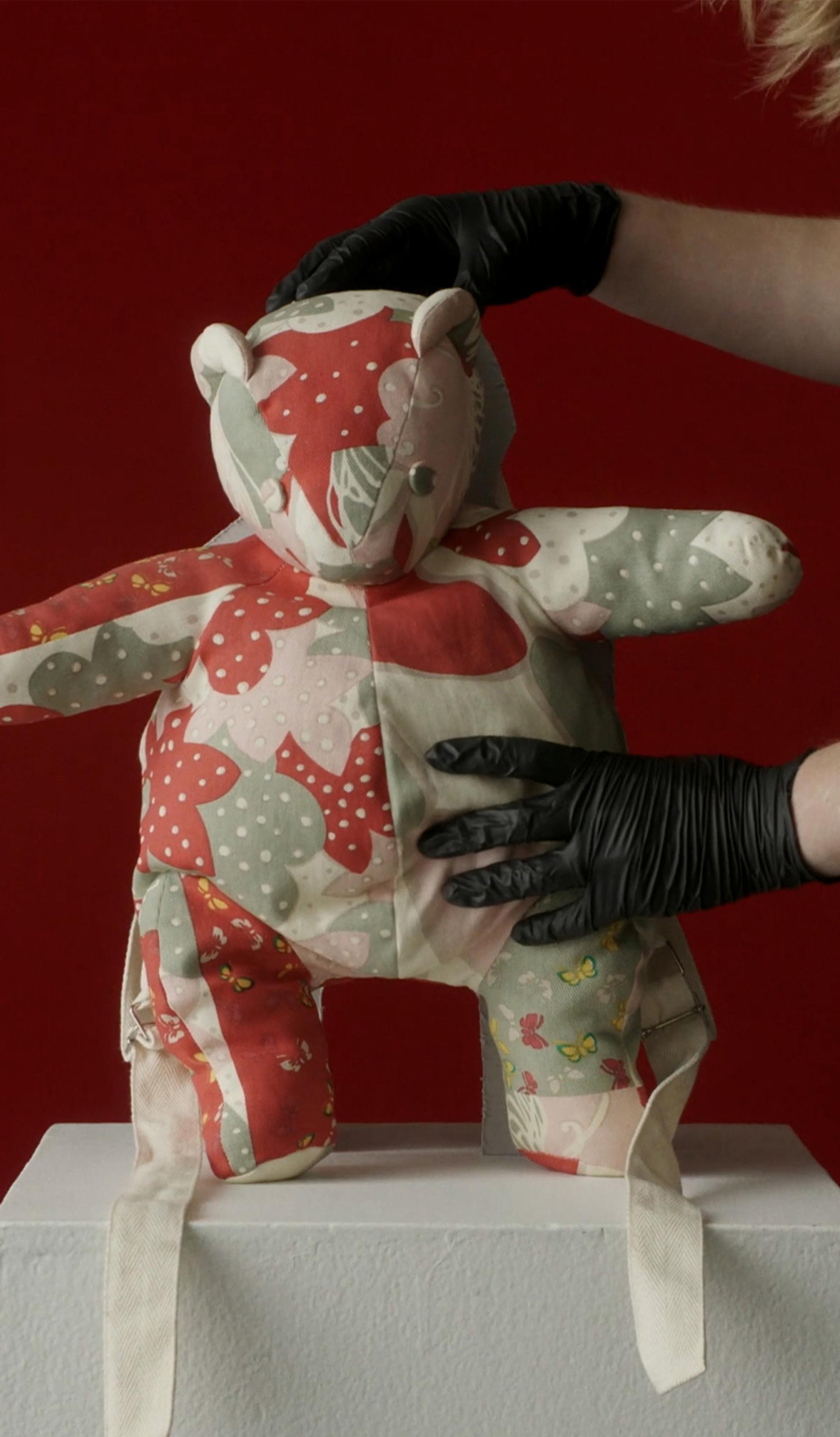 A patchwork teddy bear held by a museum curator against a red background.