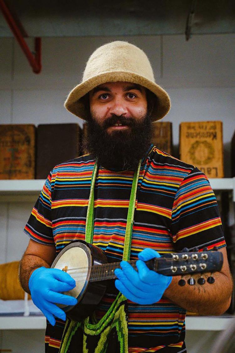 Joseph Douglas faces the camera holding a small banjo. He is wearing a yellow felt hat, striped t shirt and blue rubber gloves.