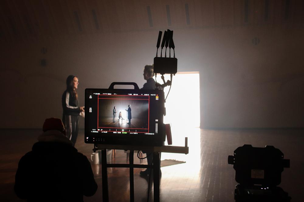 A HD filming screen captures the action of a young woman talking to a man in a large room with wooden floorboards.