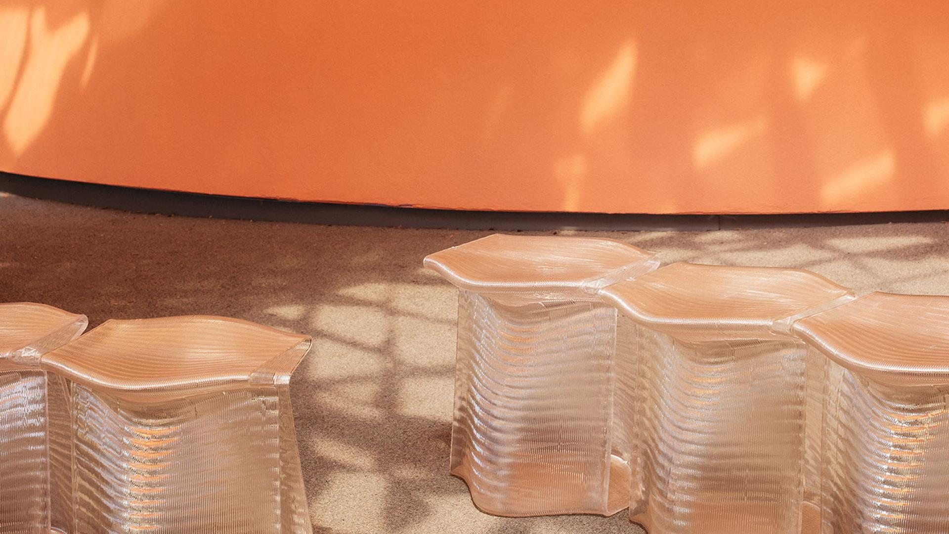 Translucent, 3D-printed stools, side-by-side, with an orange wall in the background. Stools are under dappled light cast by the canopy.