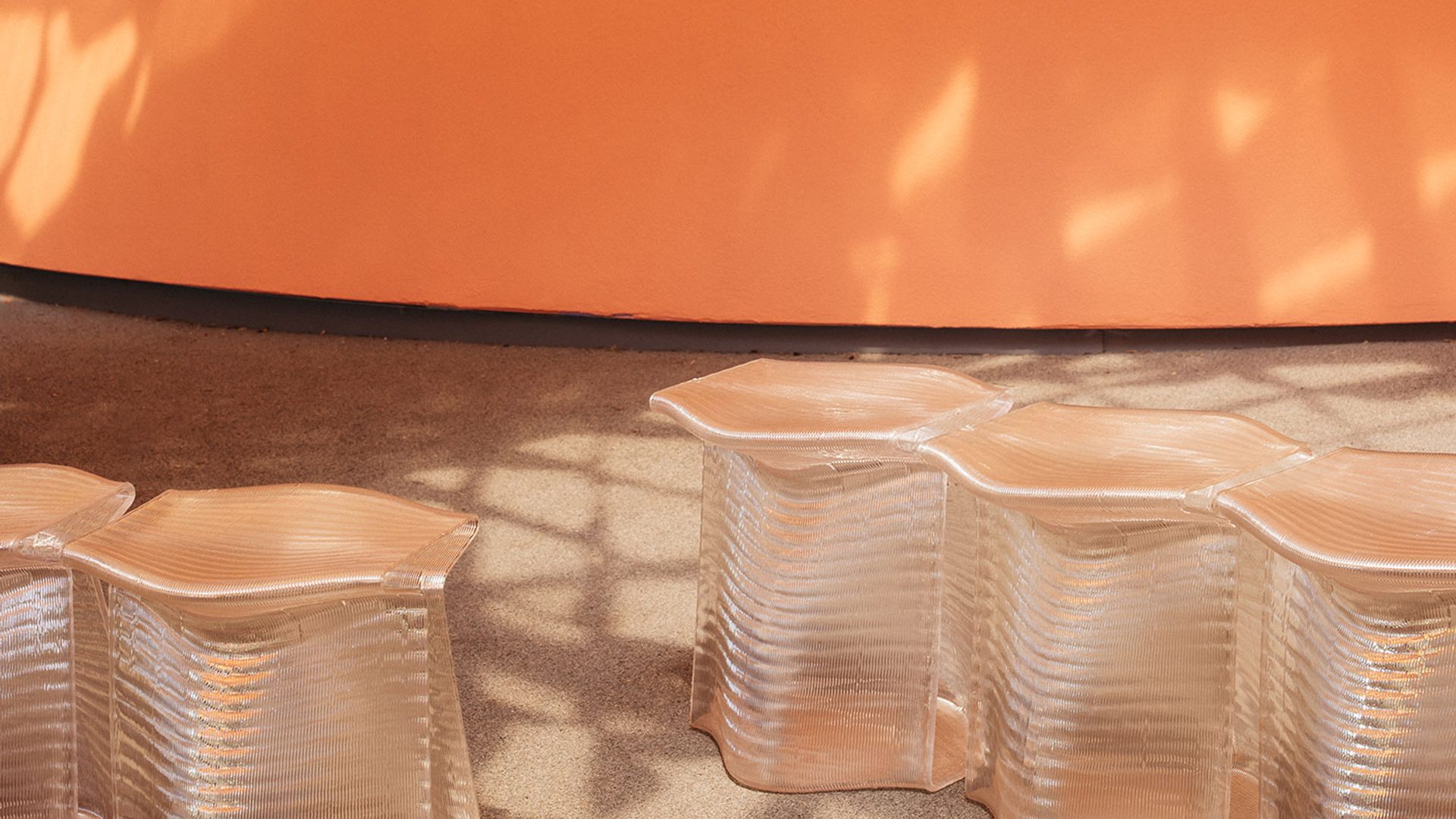 Translucent, 3D-printed stools, side-by-side, with an orange wall in the background. Stools are under dappled light cast by the canopy.