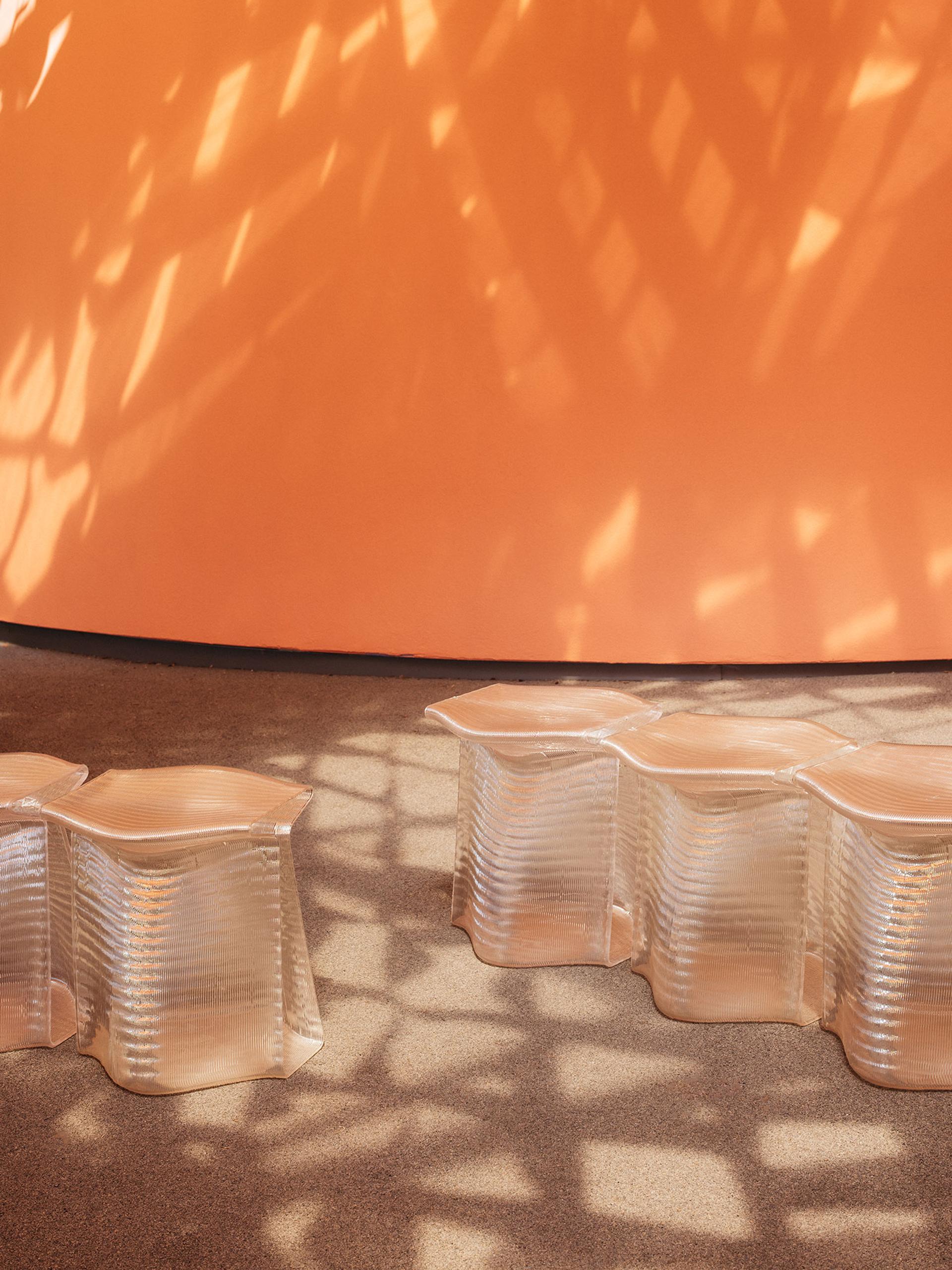 Translucent, 3D-printed stools, side-by-side, with an orange wall in the background. Stools are under dappled light cast by the canopy.