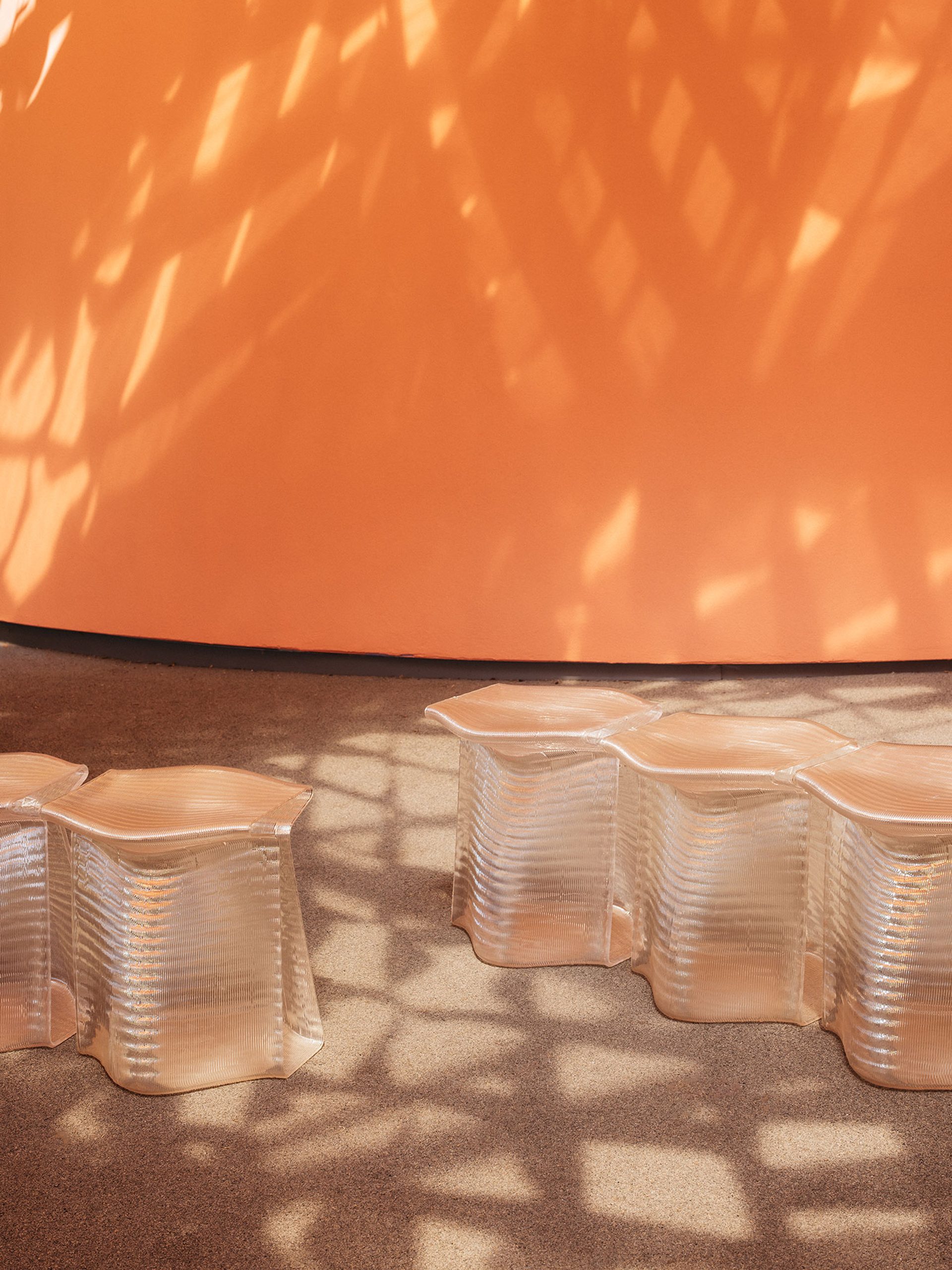 Translucent, 3D-printed stools, side-by-side, with an orange wall in the background. Stools are under dappled light cast by the canopy.