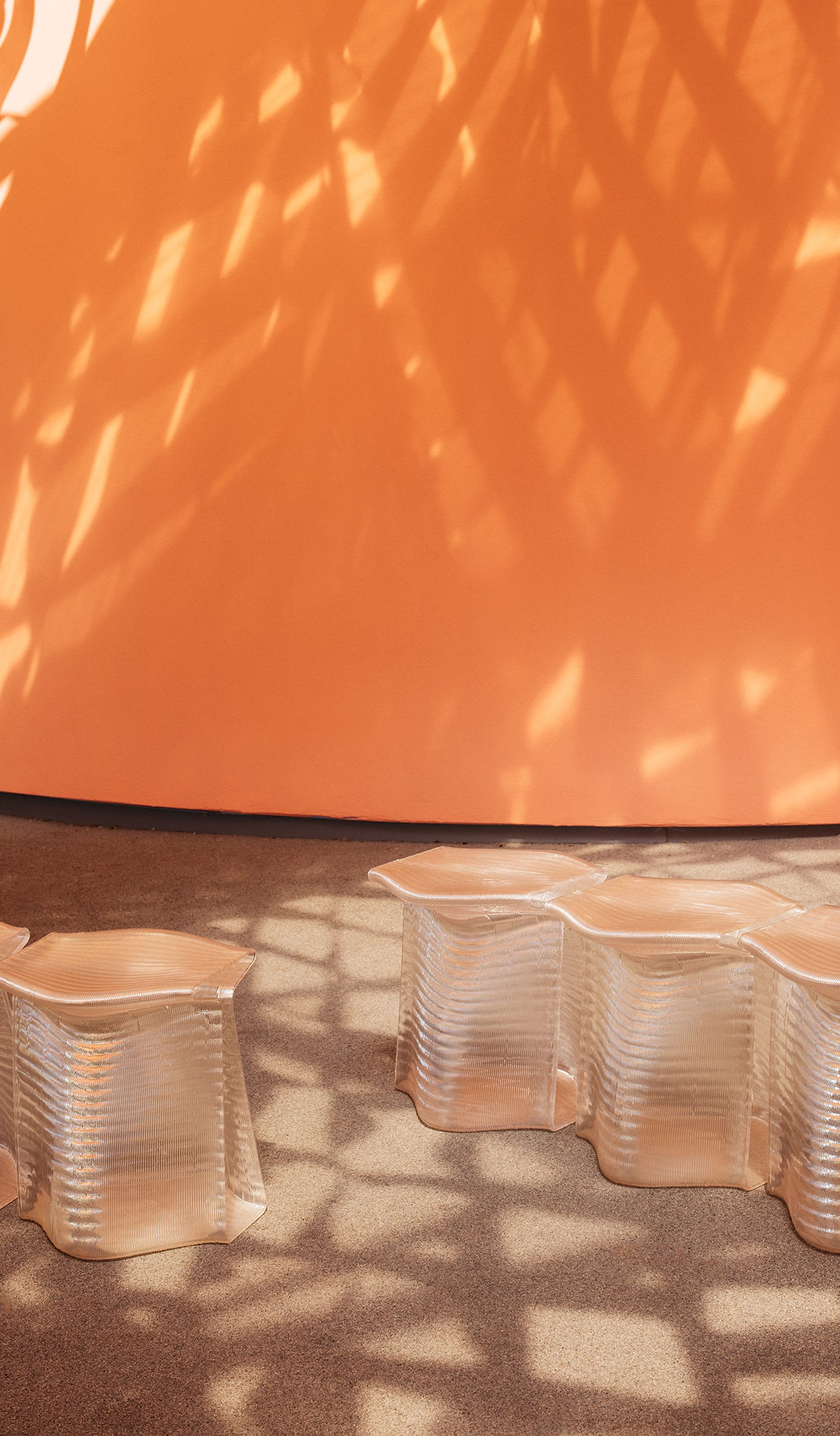 Translucent, 3D-printed stools, side-by-side, with an orange wall in the background. Stools are under dappled light cast by the canopy.