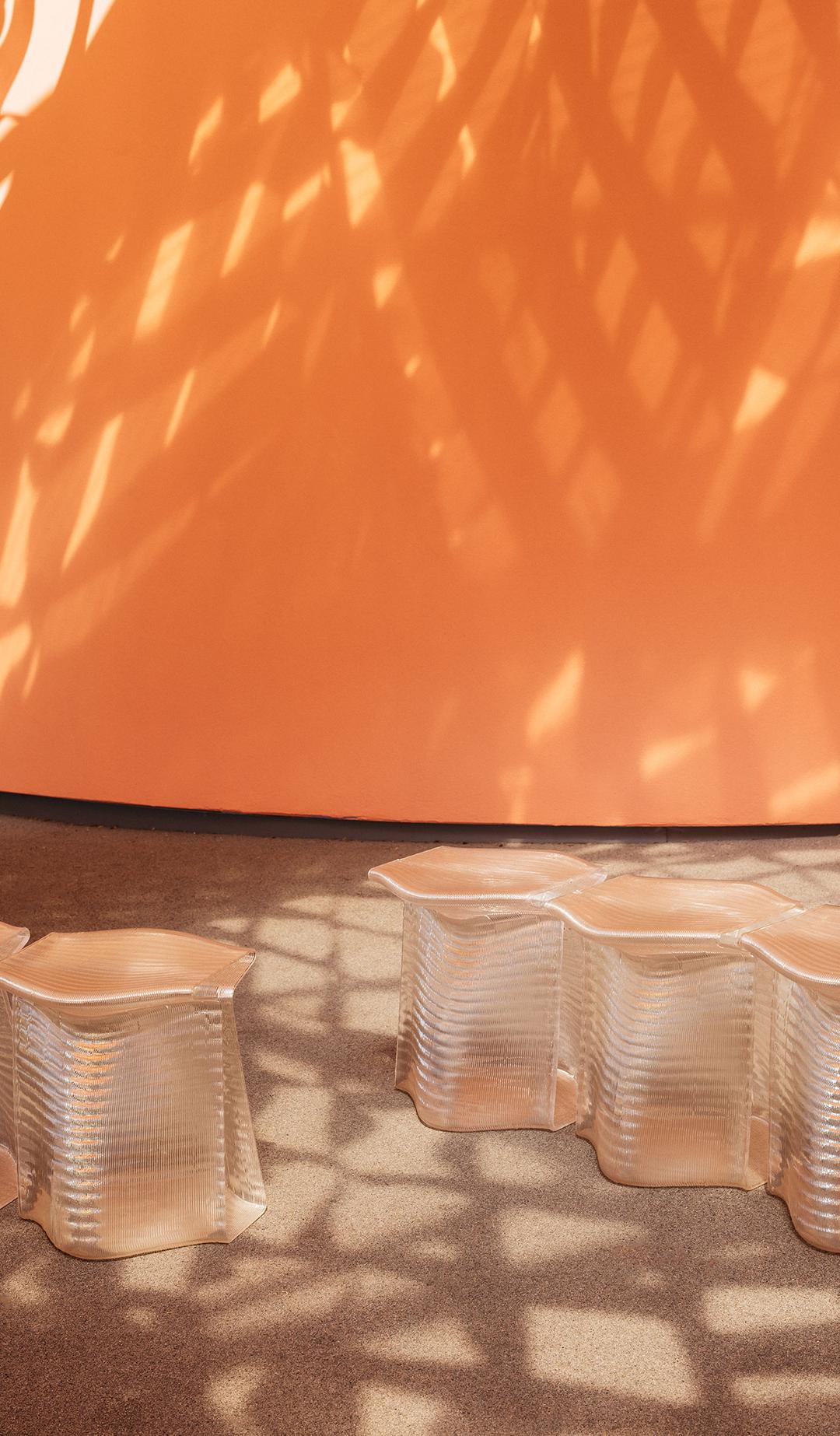 Translucent, 3D-printed stools, side-by-side, with an orange wall in the background. Stools are under dappled light cast by the canopy.