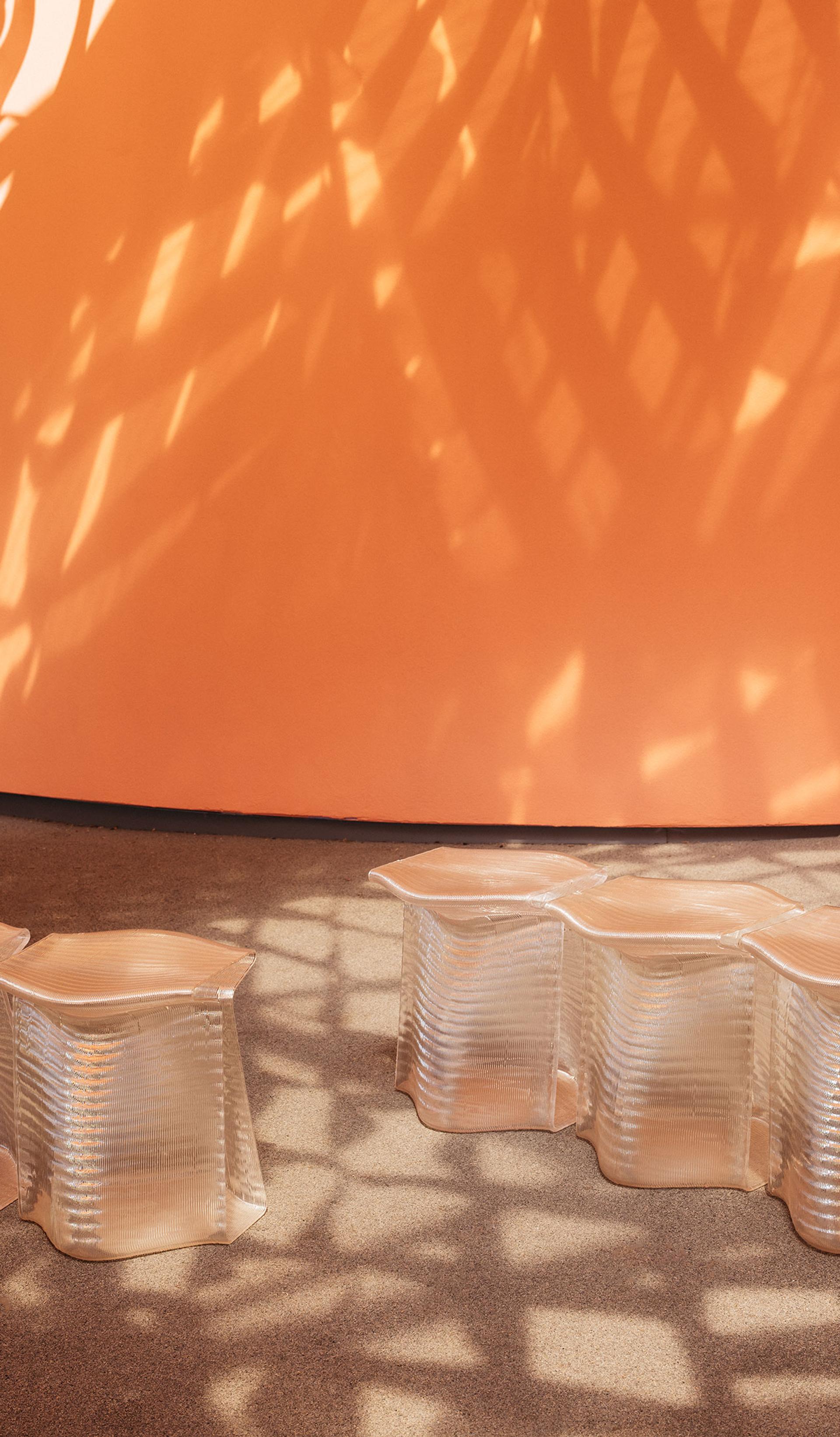 Translucent, 3D-printed stools, side-by-side, with an orange wall in the background. Stools are under dappled light cast by the canopy.