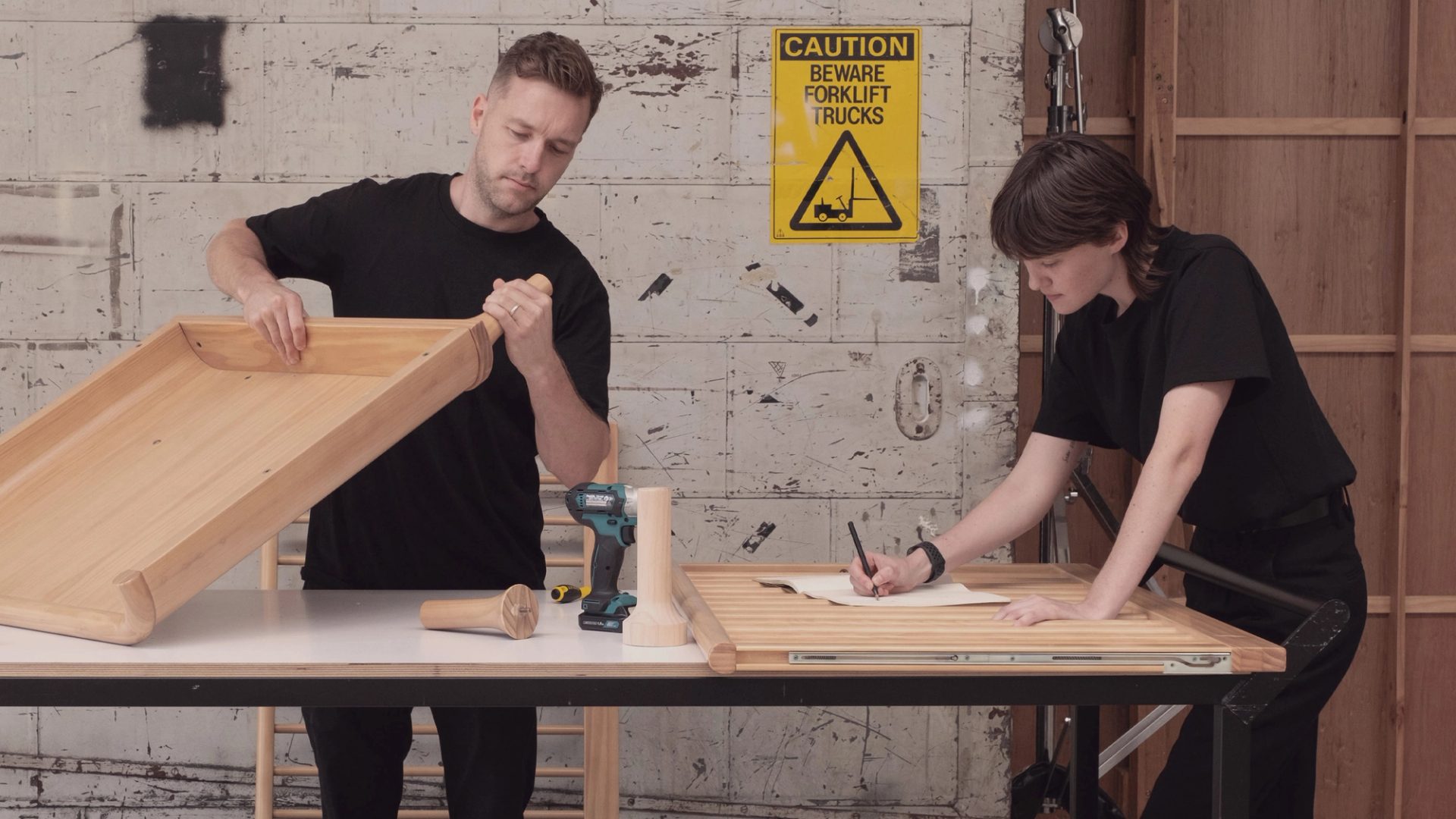 2025 Design Commission recipient Tom Skeehan holds part of his award-winning design -- a smoothly sculpted wooden cabinet -- in a workshop, alongside a fellow designer who is making notes on a sheet of paper.