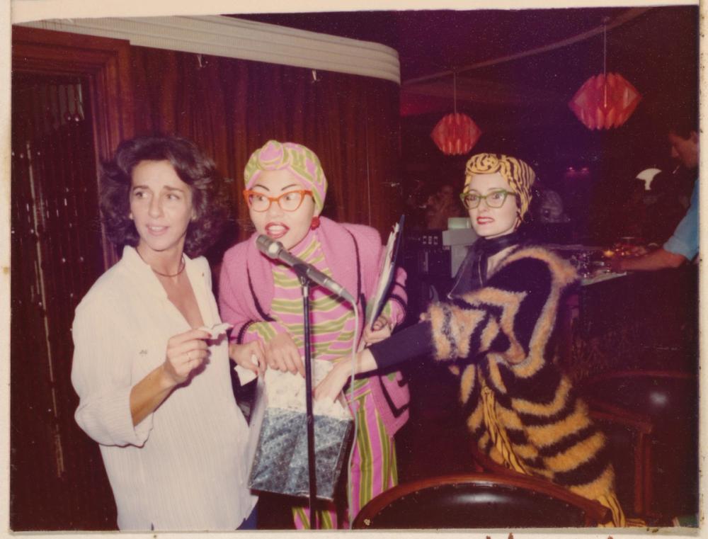 photographic print of three women in a bar standing at a microphone. the middle woman is holding a box of raffle tickets and speaking into the microphone.
