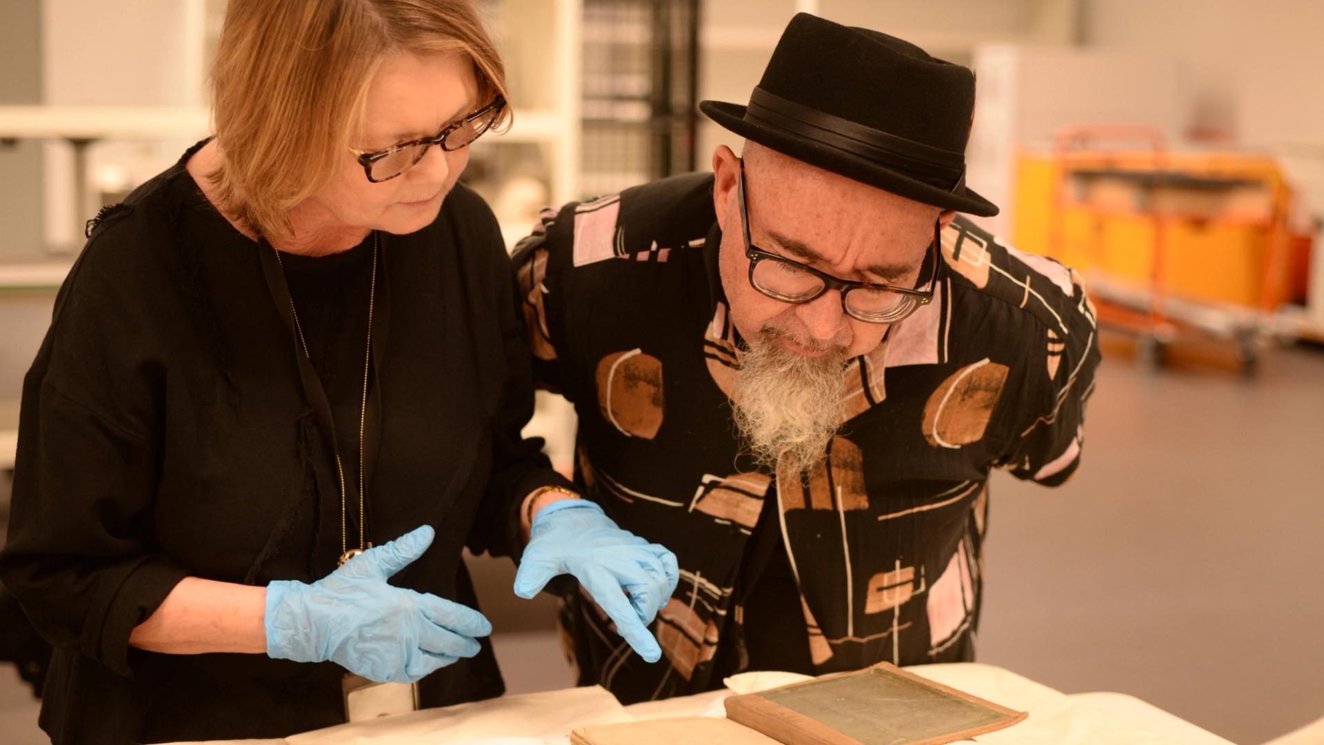 Two people leaning towards a table, wearing gloves and analysing the object on the table.