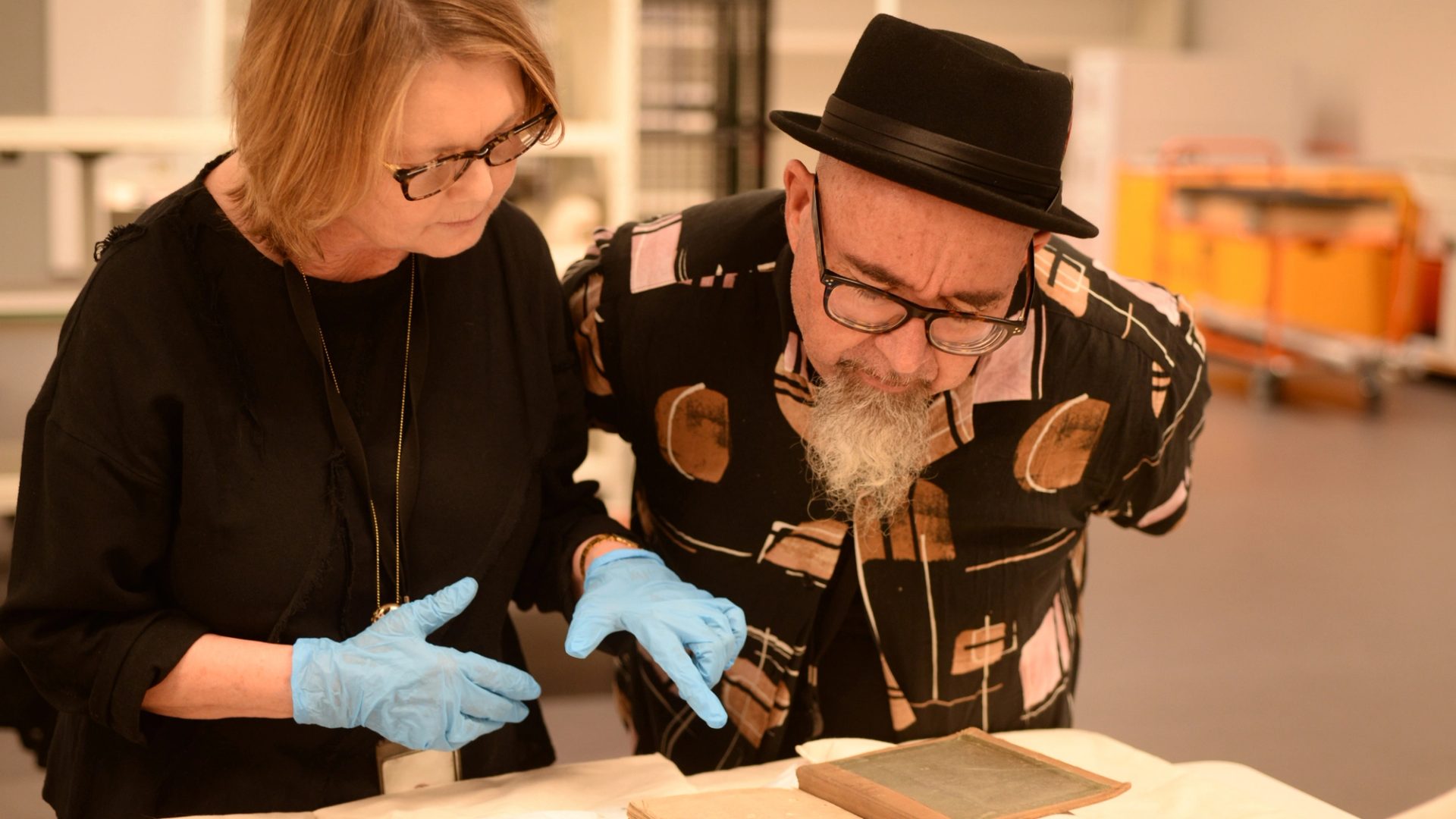 Two people leaning towards a table, wearing gloves and analysing the object on the table.