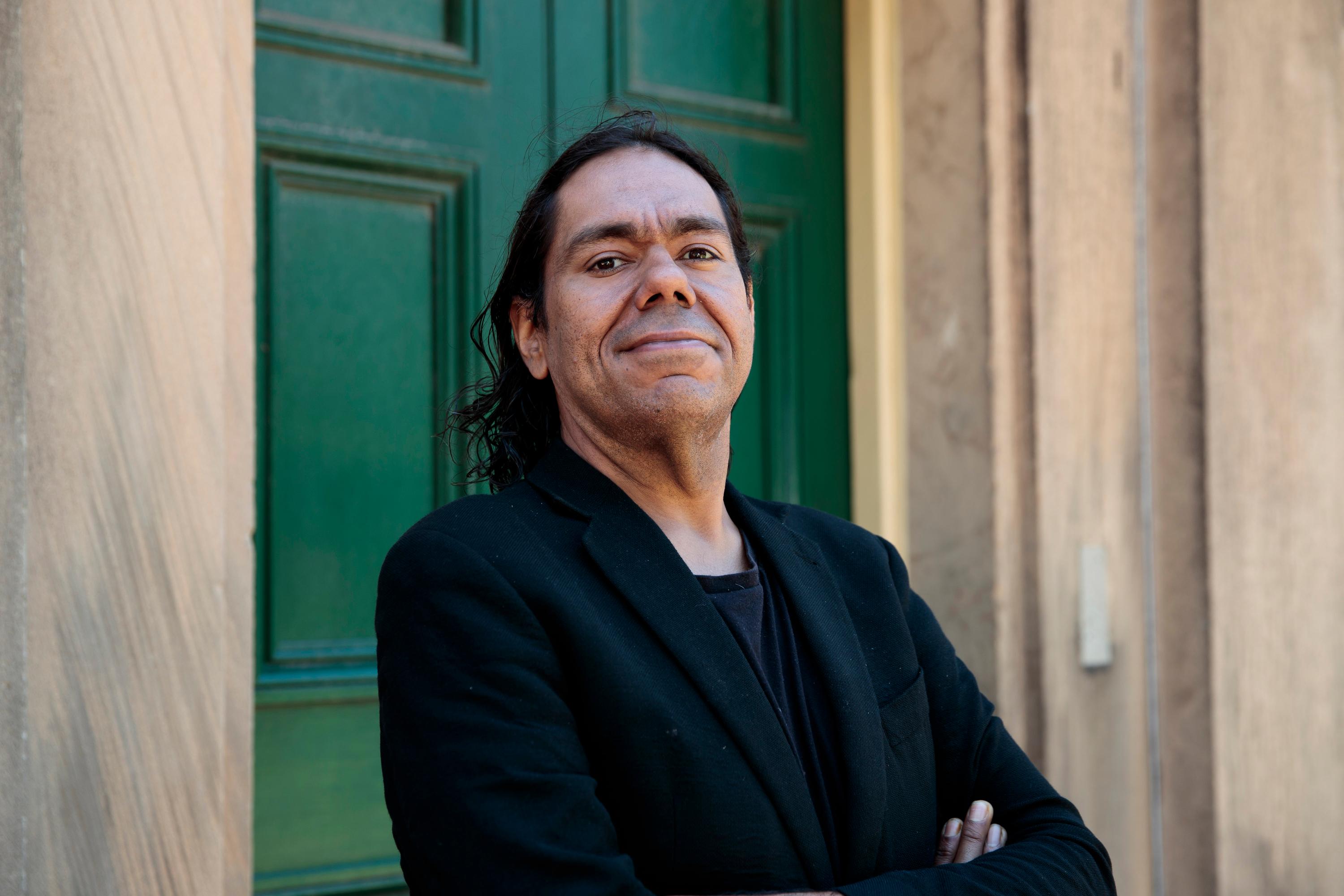 A mid shot of a man standing in front of a sandstone building with a green door. He wears a black T-shirt and black suit jacket. His arms are crossed in front of his body. His shoulder length black hair is pushed behind him, and he is smiling with his mouth closed.