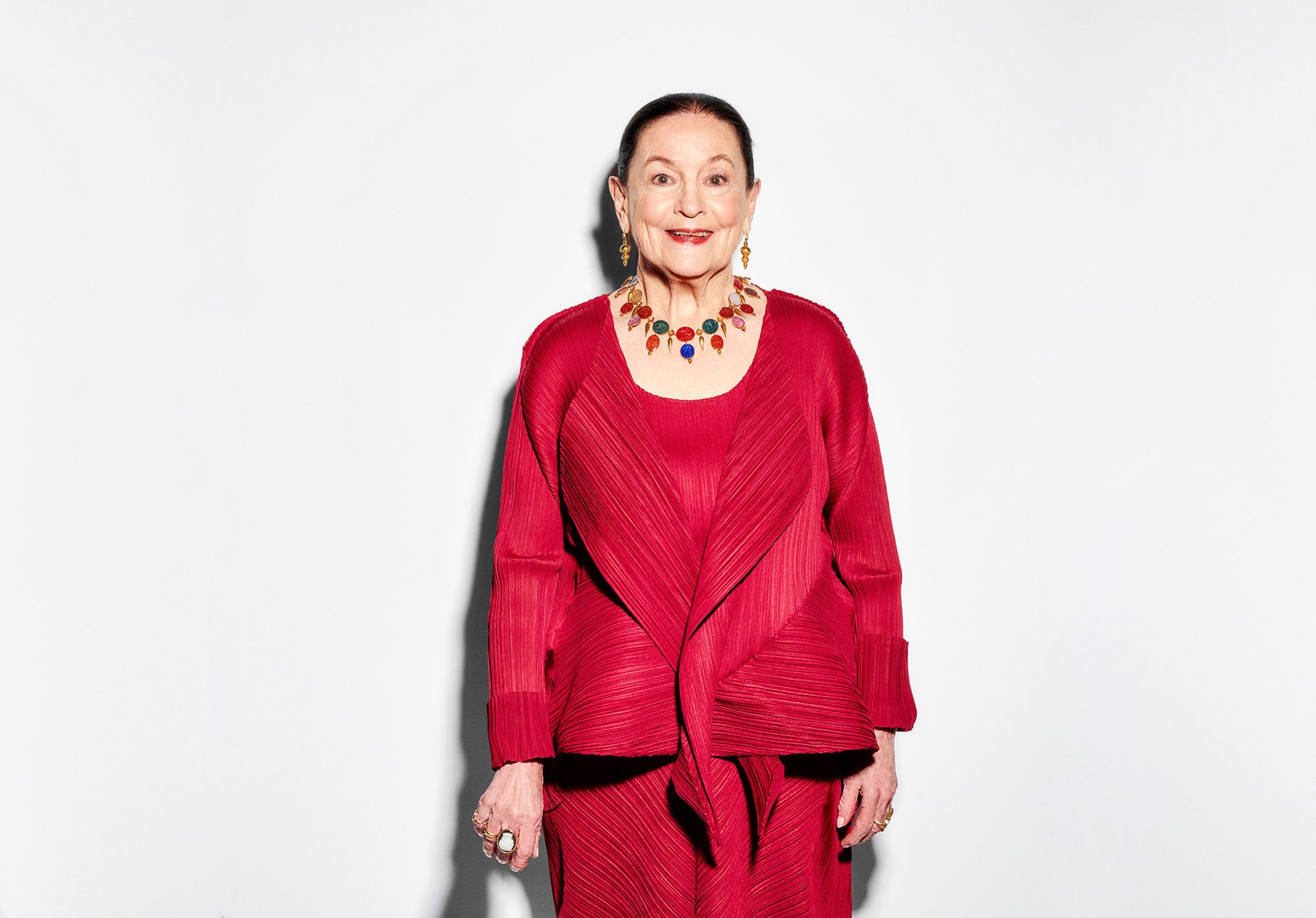 A dark haired woman wearing a red three piece outfit and a gold necklace set with multi-coloured stones that have faces carved on them.