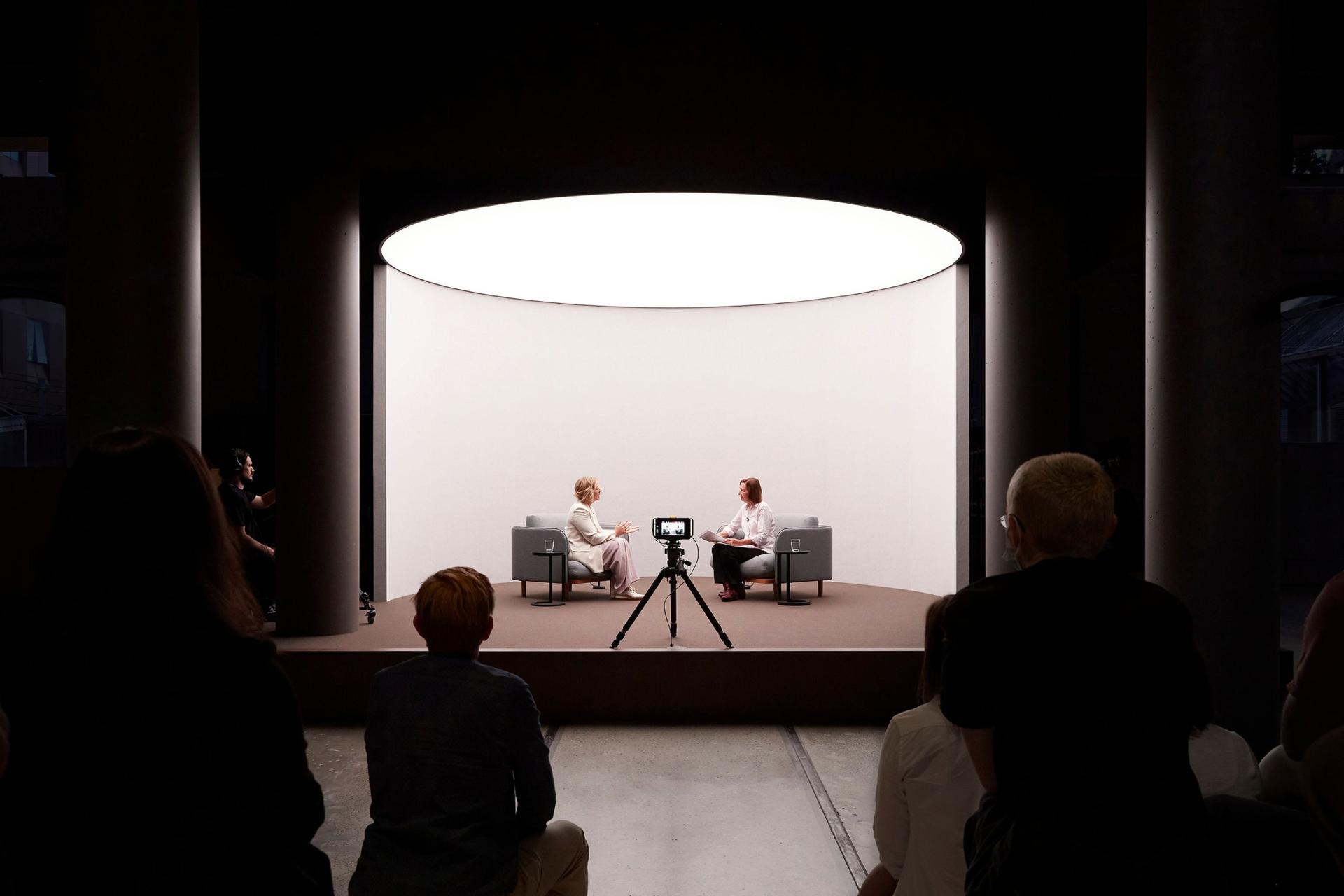 Two women speaking on a short stage in front of a camera and an audience.