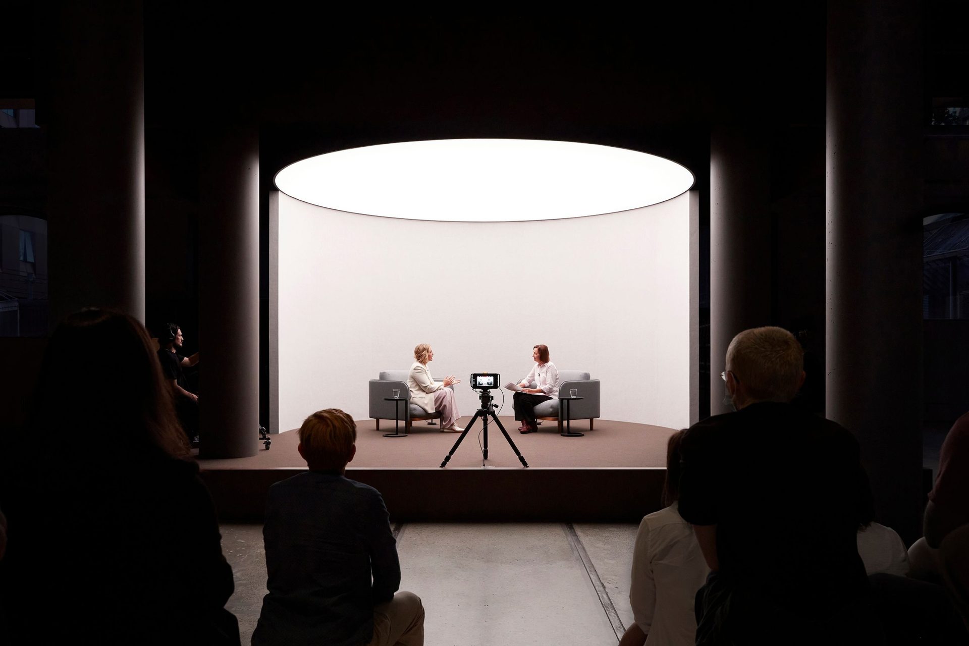 two women speaking on a short stage in front of a camera and an audience