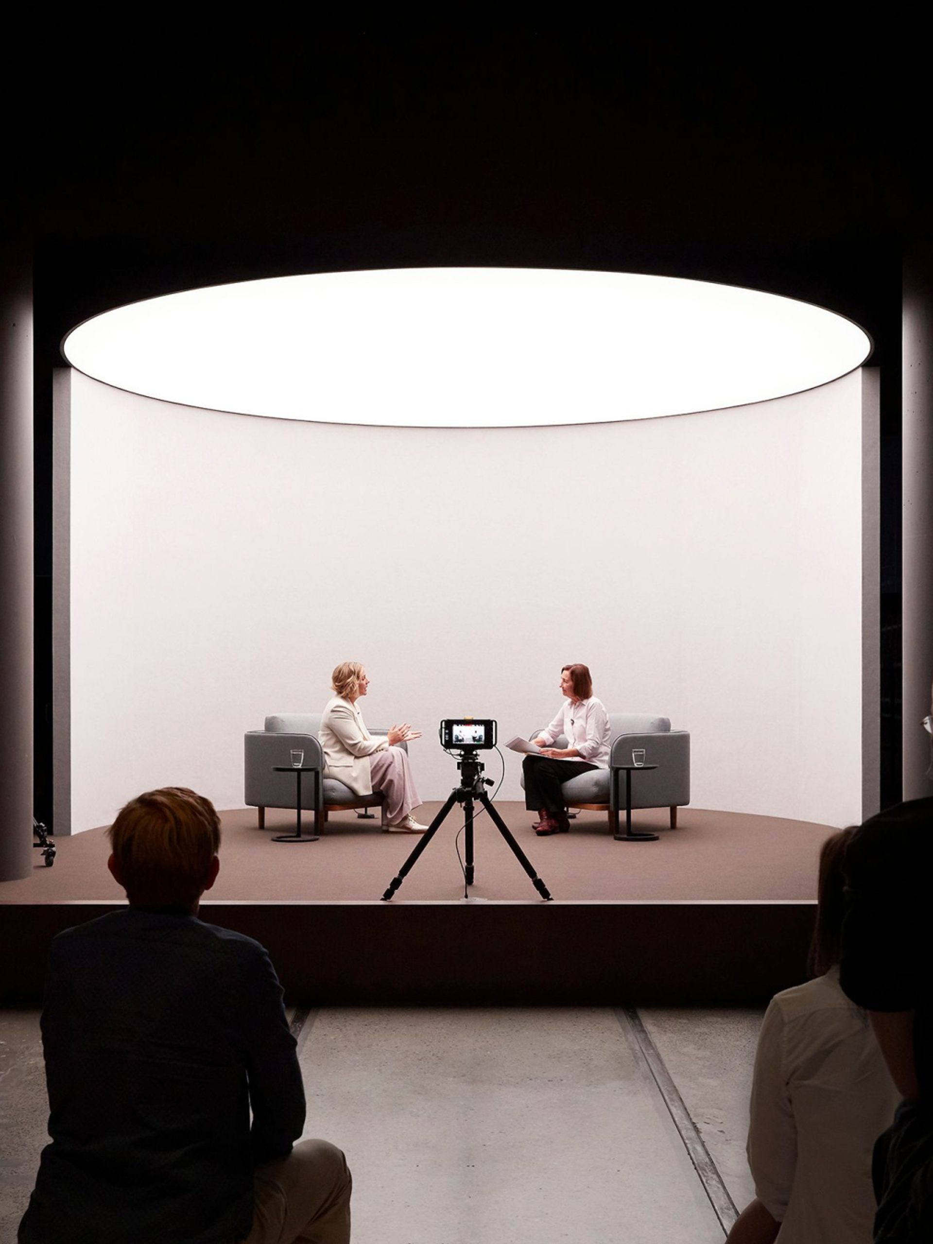 two women speaking on a short stage in front of a camera and an audience