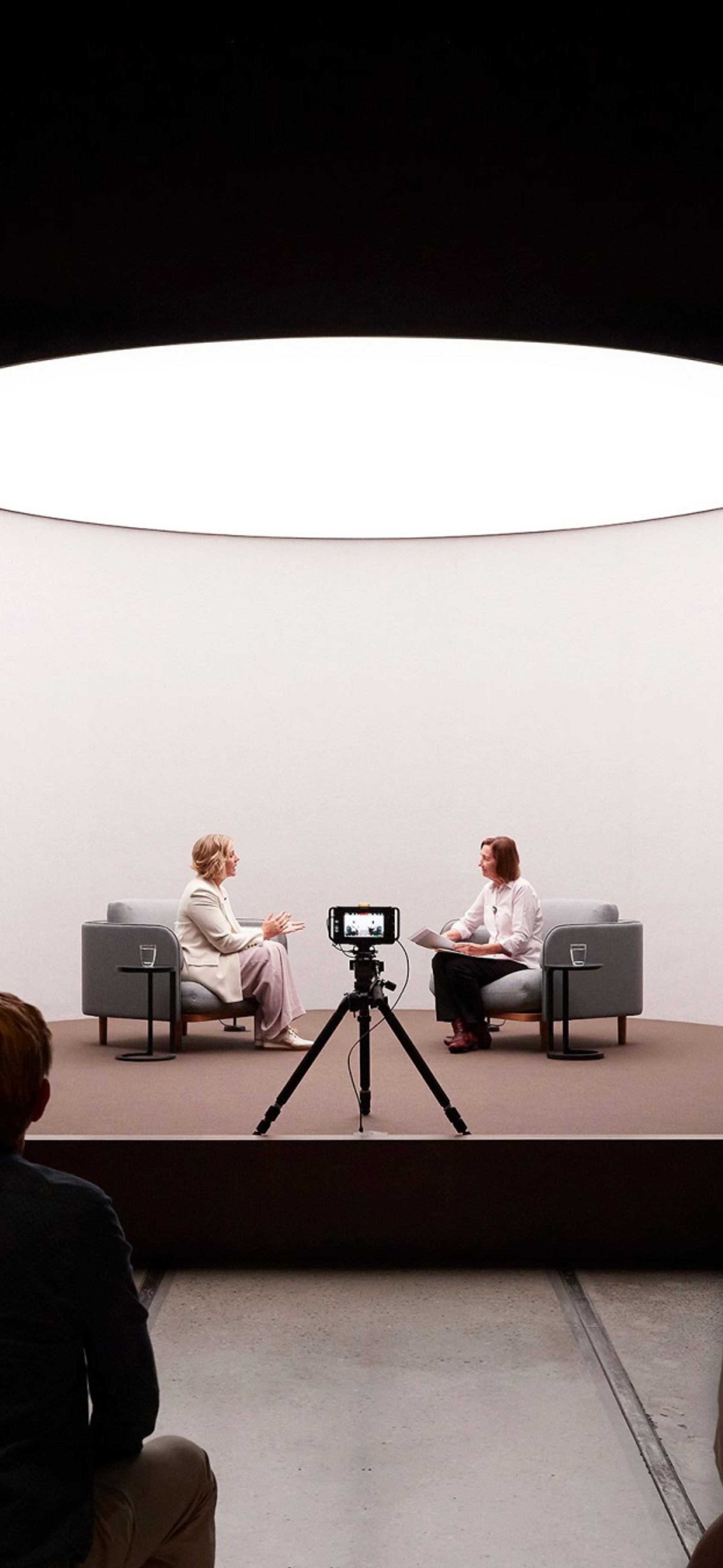 two women speaking on a short stage in front of a camera and an audience