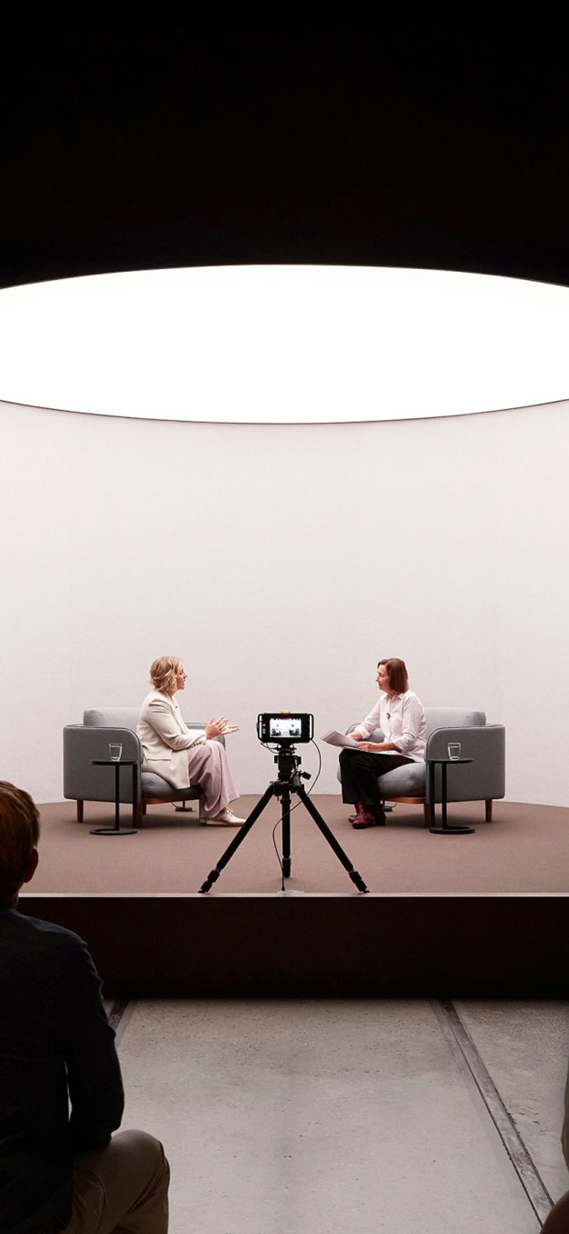 Two women speaking on a short stage in front of a camera and an audience.