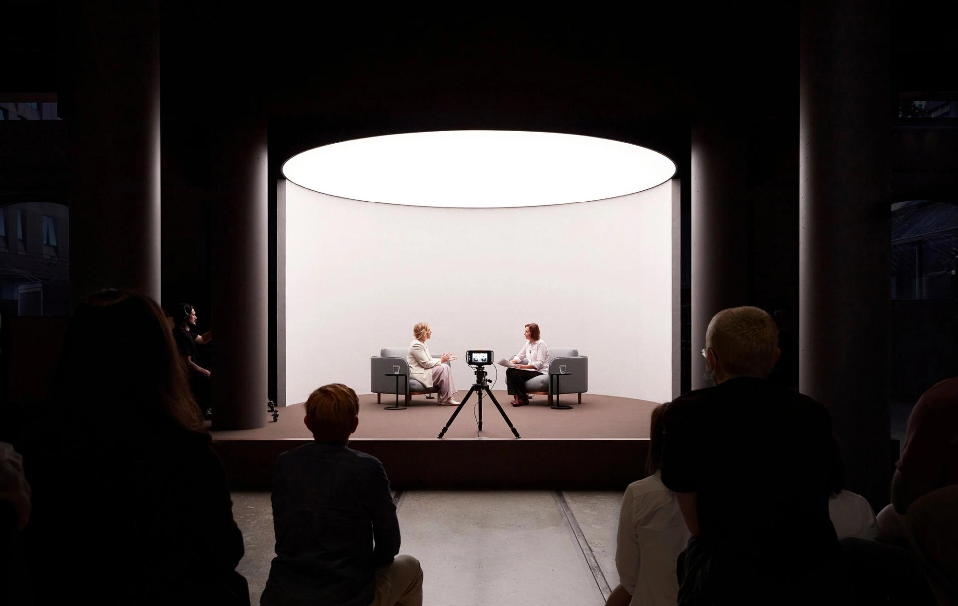 Two women speaking on a short stage in front of a camera and an audience.