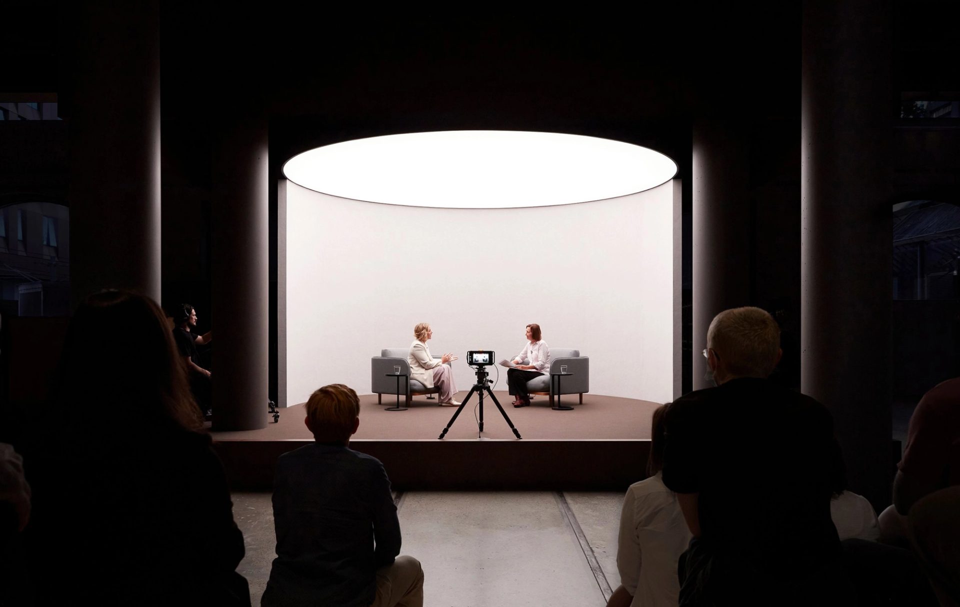 two women speaking on a short stage in front of a camera and an audience