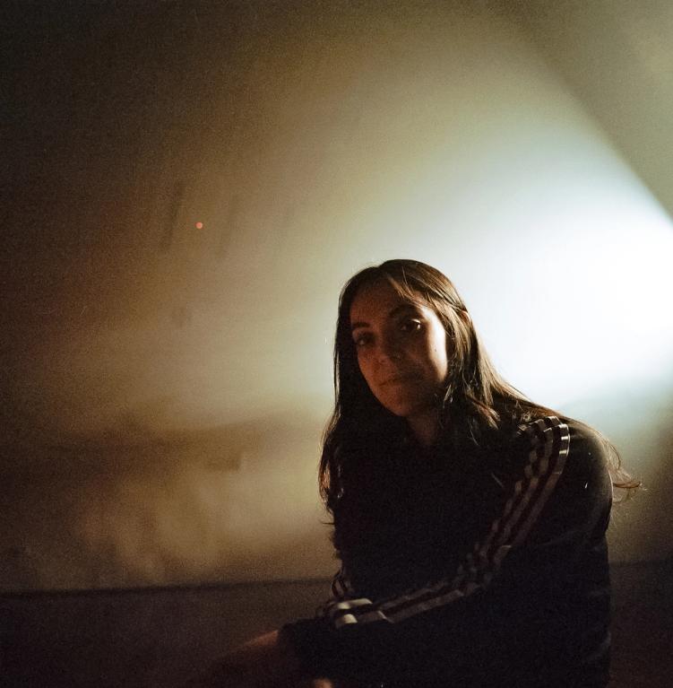 A young woman stands in the middle of a dark room. She has long dark hair and is wearing a black Adidas jacket with white stripes. She smiles softly.