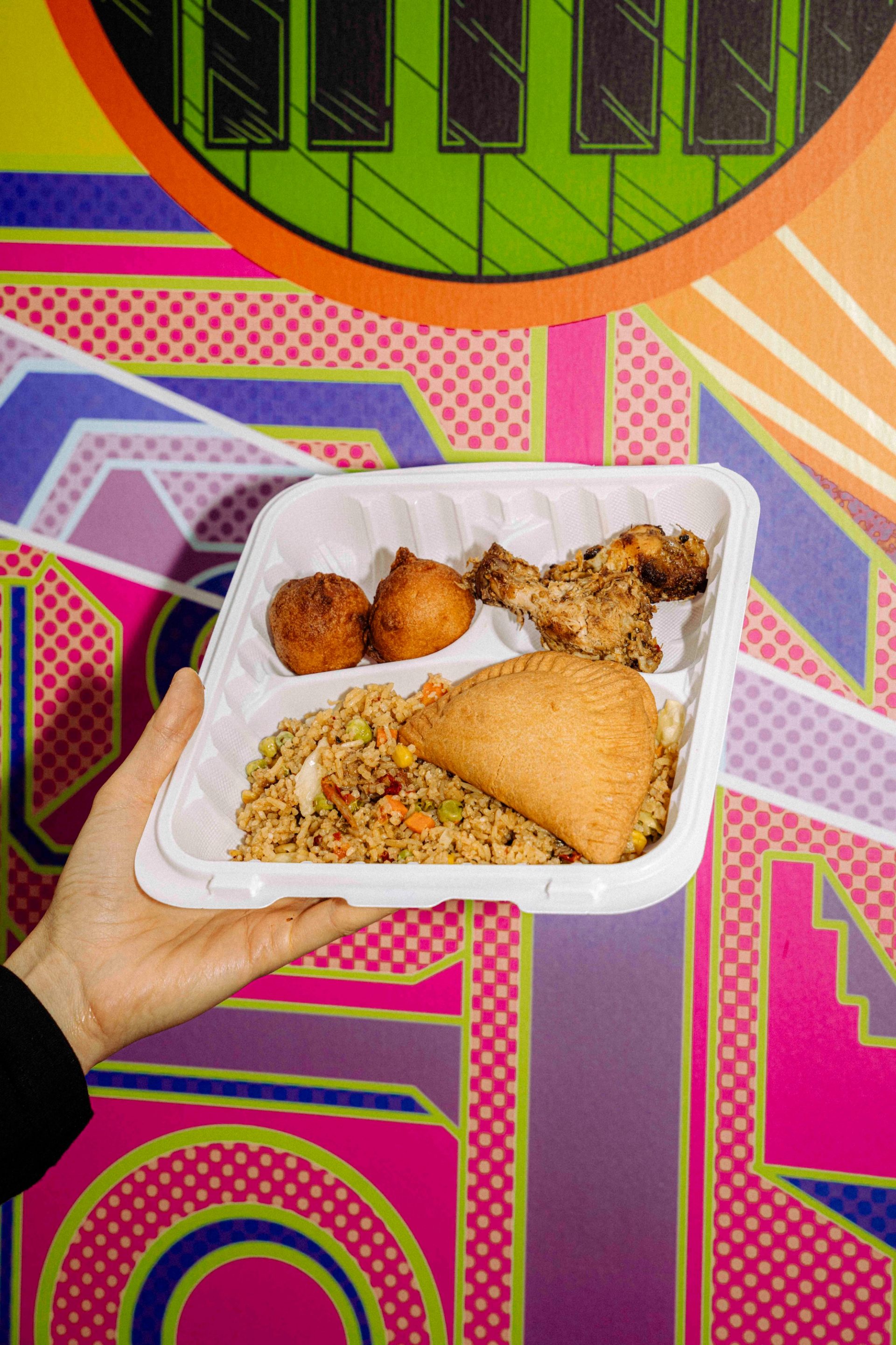 A hand holds a white tray filled with various foods, including a rice dish, a pastry, two fried balls and a piece of meat, against a colourful geometric mural.