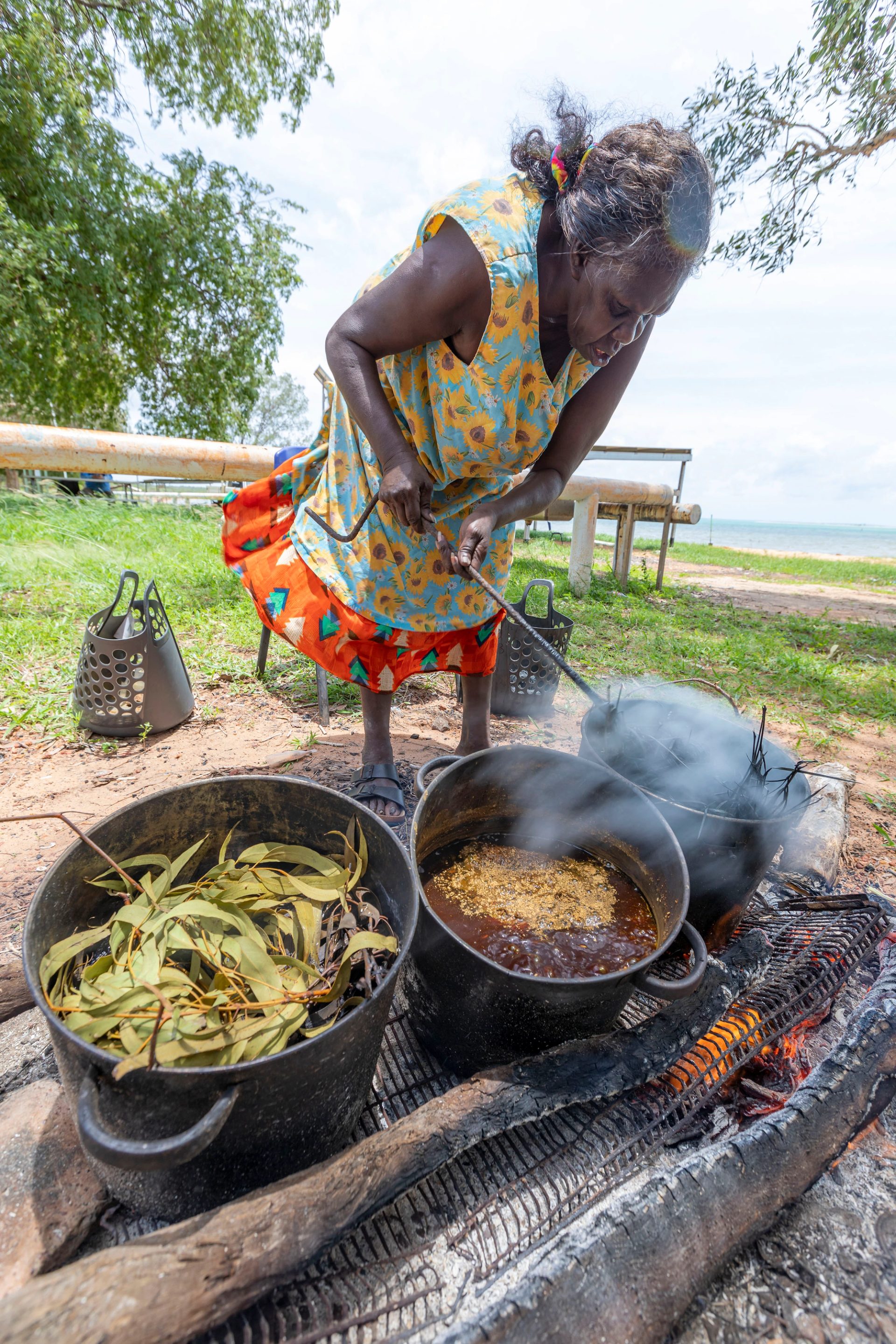 A woman in a brightly coloured dress stands outside and is leaning over three large pots filled with different leaves and plants. She is poking the pot on the far left with a stick. The sky is blue, and the ocean is visible in the background.