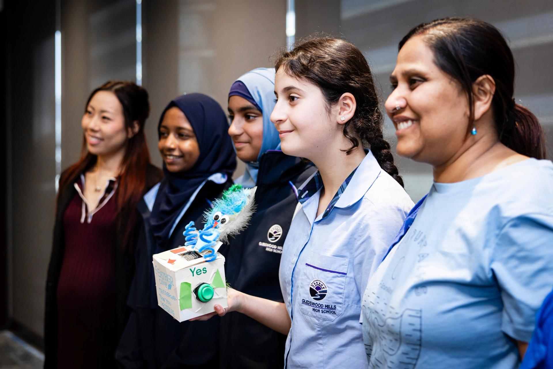 Group photo of students and teachers smiling and stood beside each other, the student in the centre holds a hand crafted mechanical device.